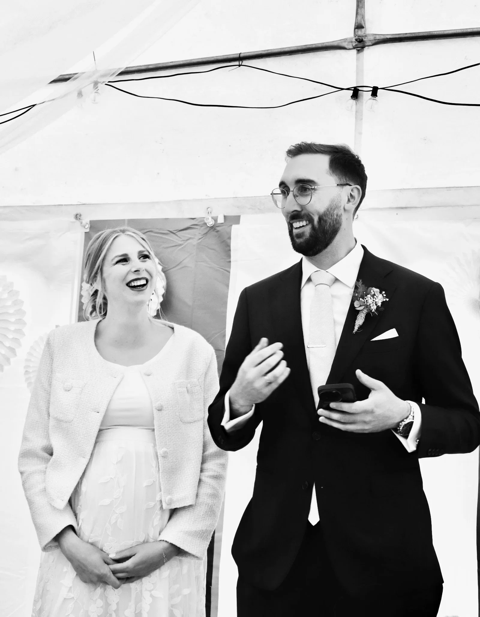 Lachendes Hochzeitspaar unter einem Festzelt; der Bräutigam hält ein Smartphone, während er eine lustige Rede hält