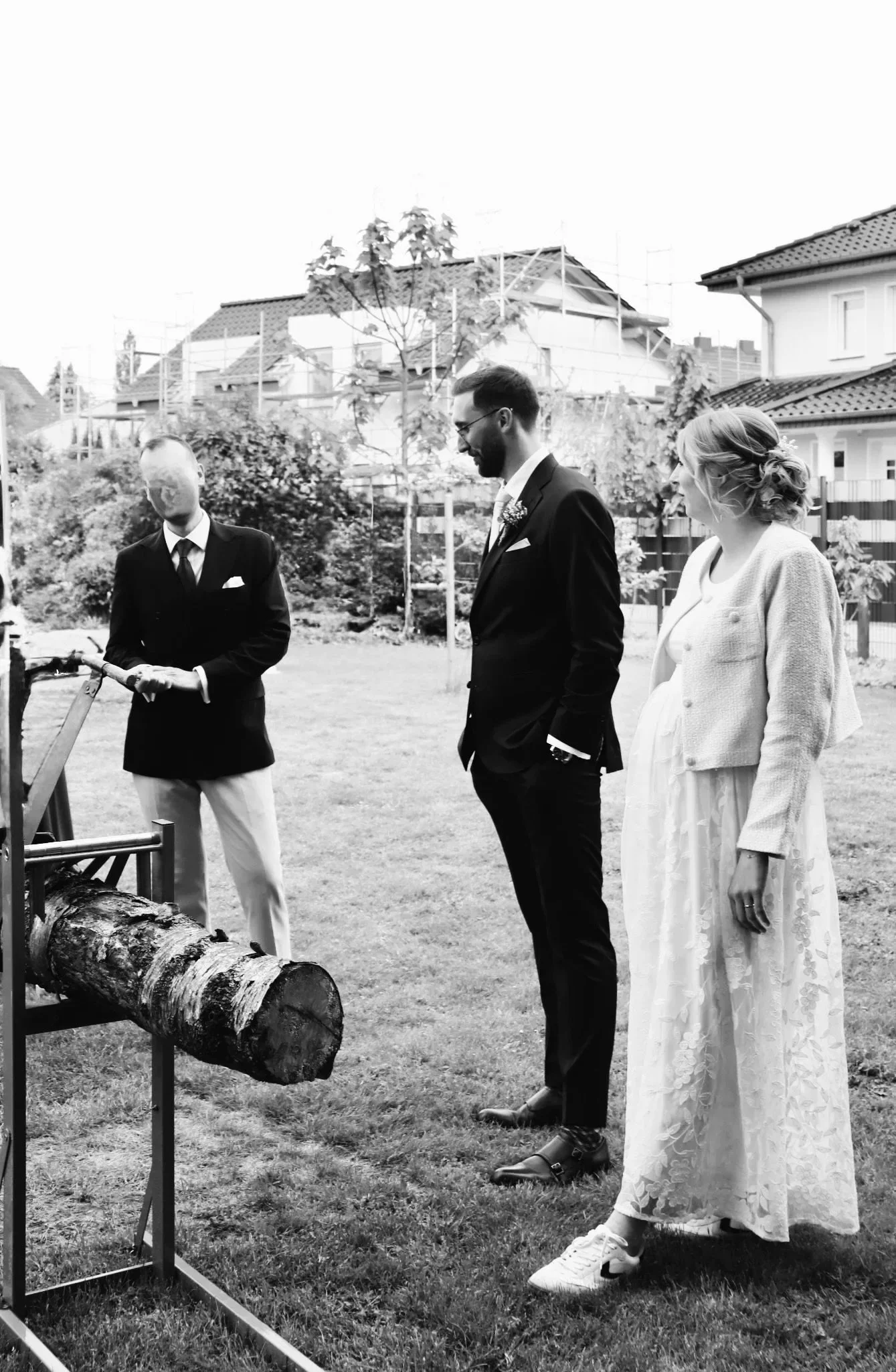 Schwarzweißfoto einer Hochzeitsfeier im Freien: Braut und Bräutigam stehen neben einem Baumstamm-Schneidegerät, begleitet von einem Mann im Anzug.