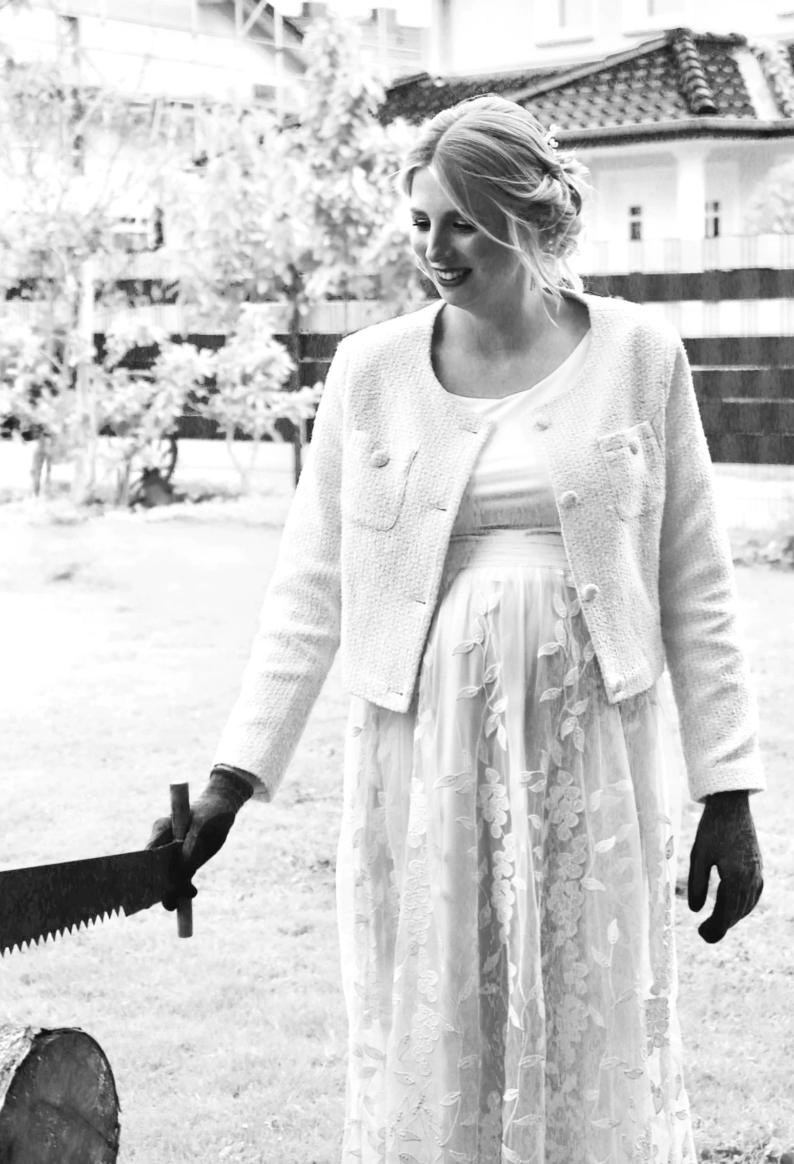 Schwarzweißfoto einer lächelnden, schwangeren Braut in elegantem Hochzeitskleid, Strickjacke und schwarzen Handschuhen, die im Garten eine große Säge hält.