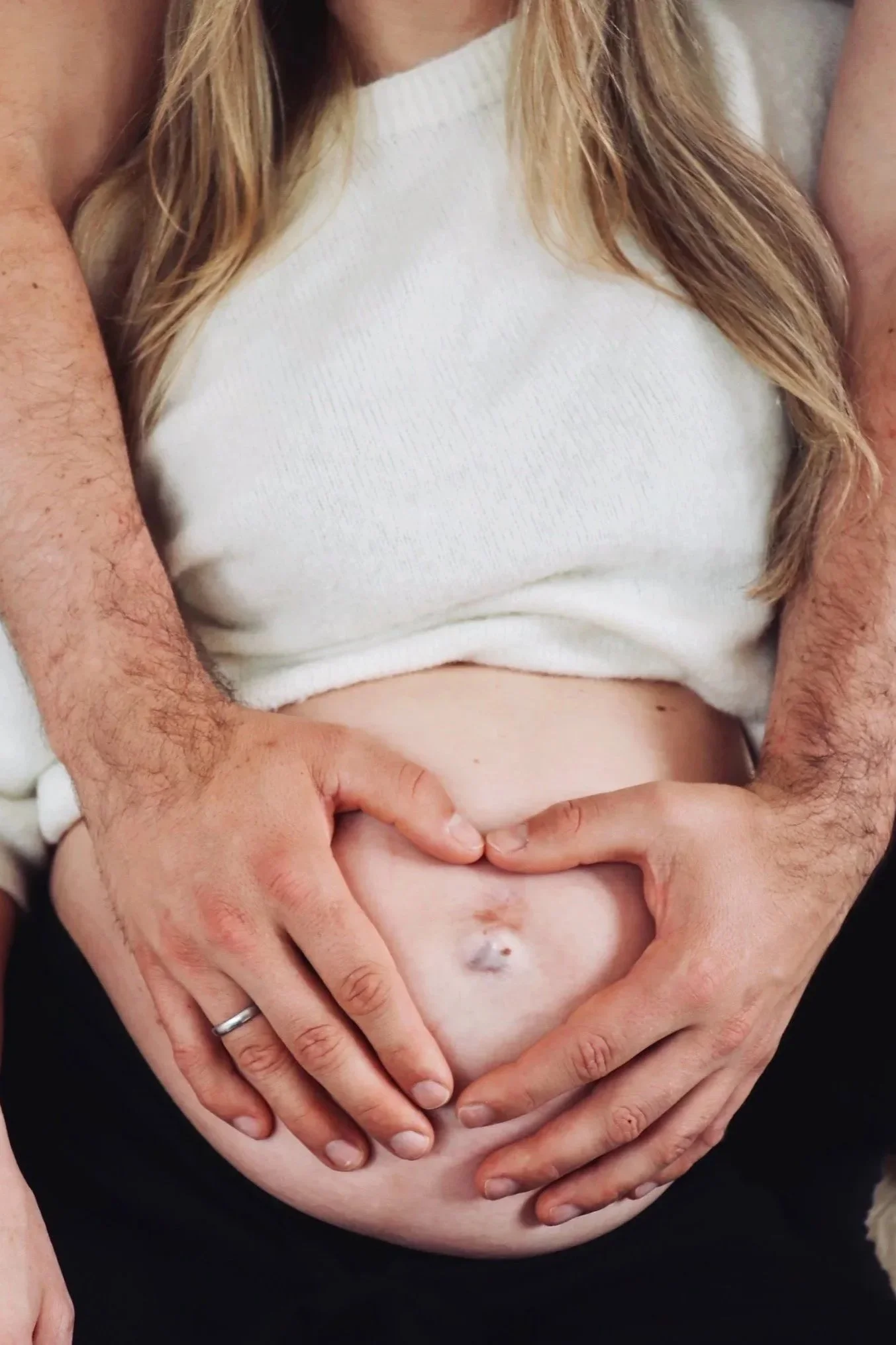 Schwangerschaftsfotografie Nähe Hannover, Bremen, Nienburg und Region,Schwangere Person mit sichtbarem Bauchnabel, deren Bauch von den Händen des Partners umschlossen wird, welche ein Herz formen