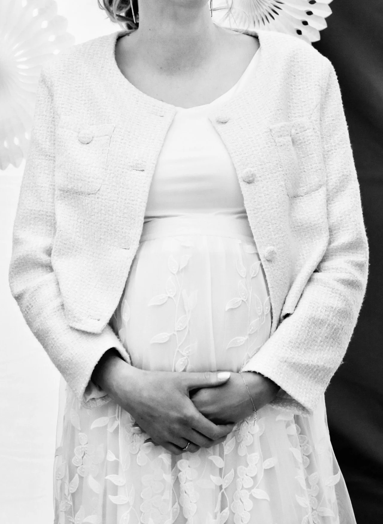 Schwarzweißaufnahme einer Frau in weißem Hochzeitskleid und strukturierter Jacke mit gefalteten Händen vor dem schwangeren Babybauch