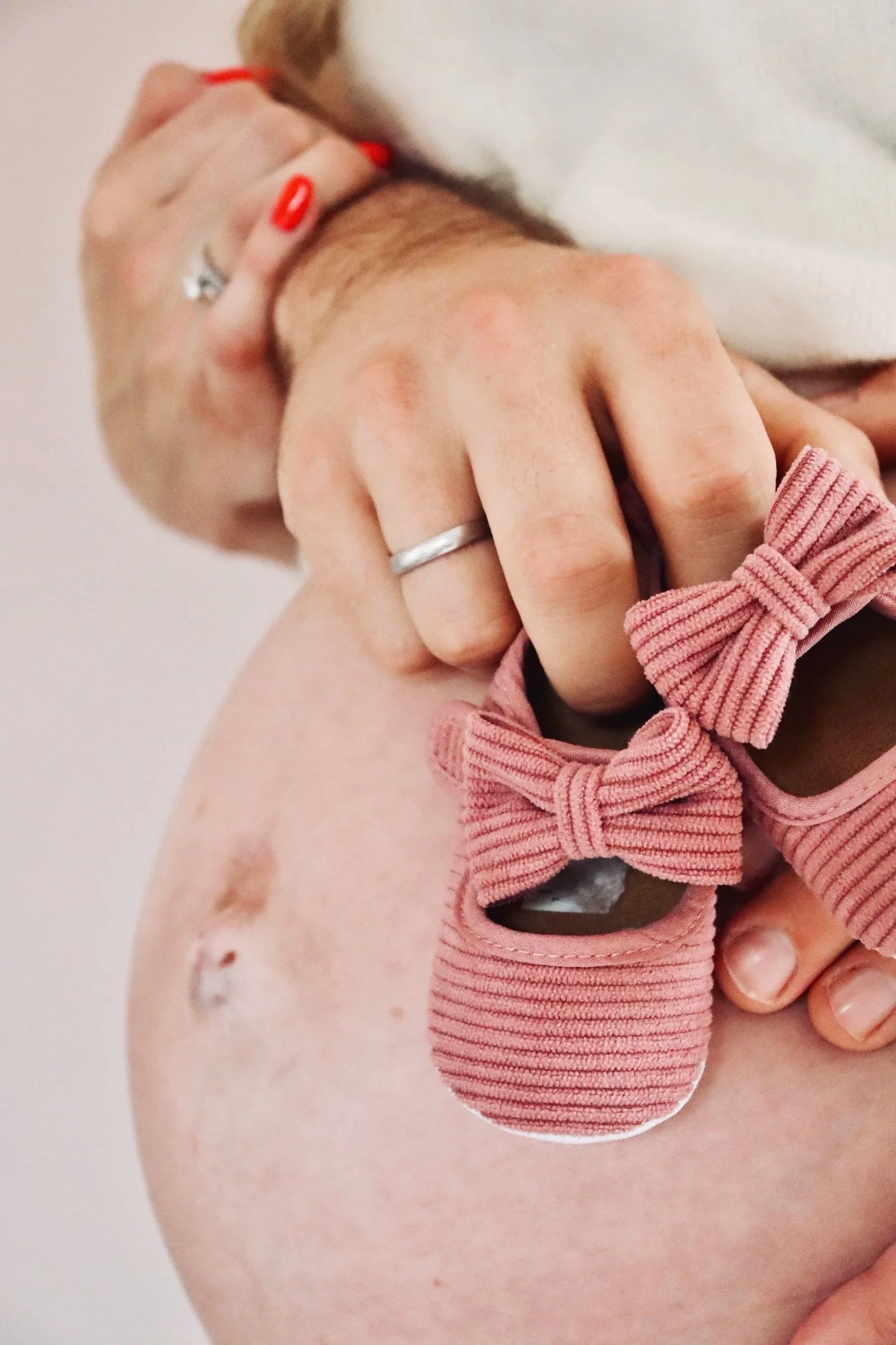 Schwangerschaftsfotografie Nähe Hannover, Bremen, Nienburg und Region, Nahaufnahme eines Babybauches mit einem rosa Strickschuh für Babys mit einer Schleife, gehalten von einer erwachsenen Hand mit Ring und rotem Nagellack.