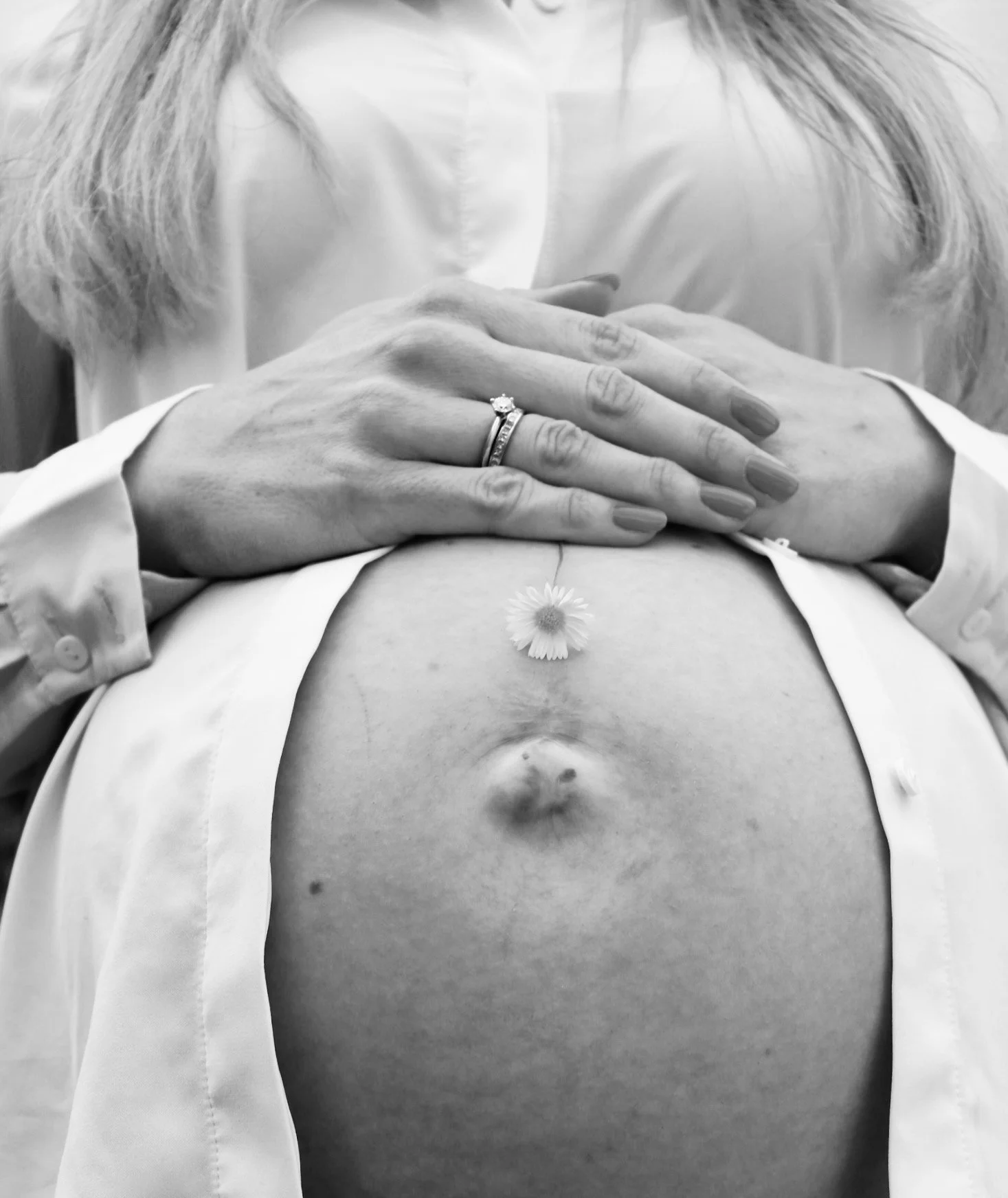 Schwarzweißfoto einer schwangeren Frau mit gefalteten Händen, Ehering und kleiner Blume auf dem Bauch