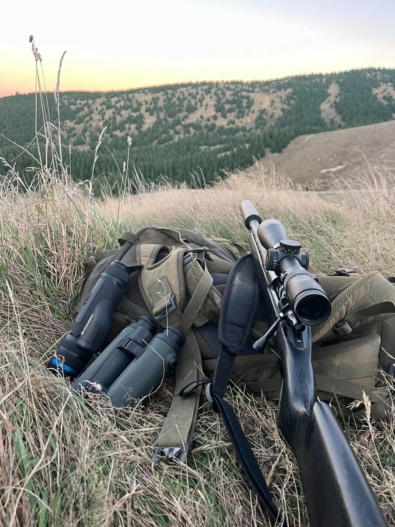 A pair of binoculars, PULSAR handheld thermal, a backpack, and a rifle resting on a grassy hillside with a view of forested rolling hills at sunset.