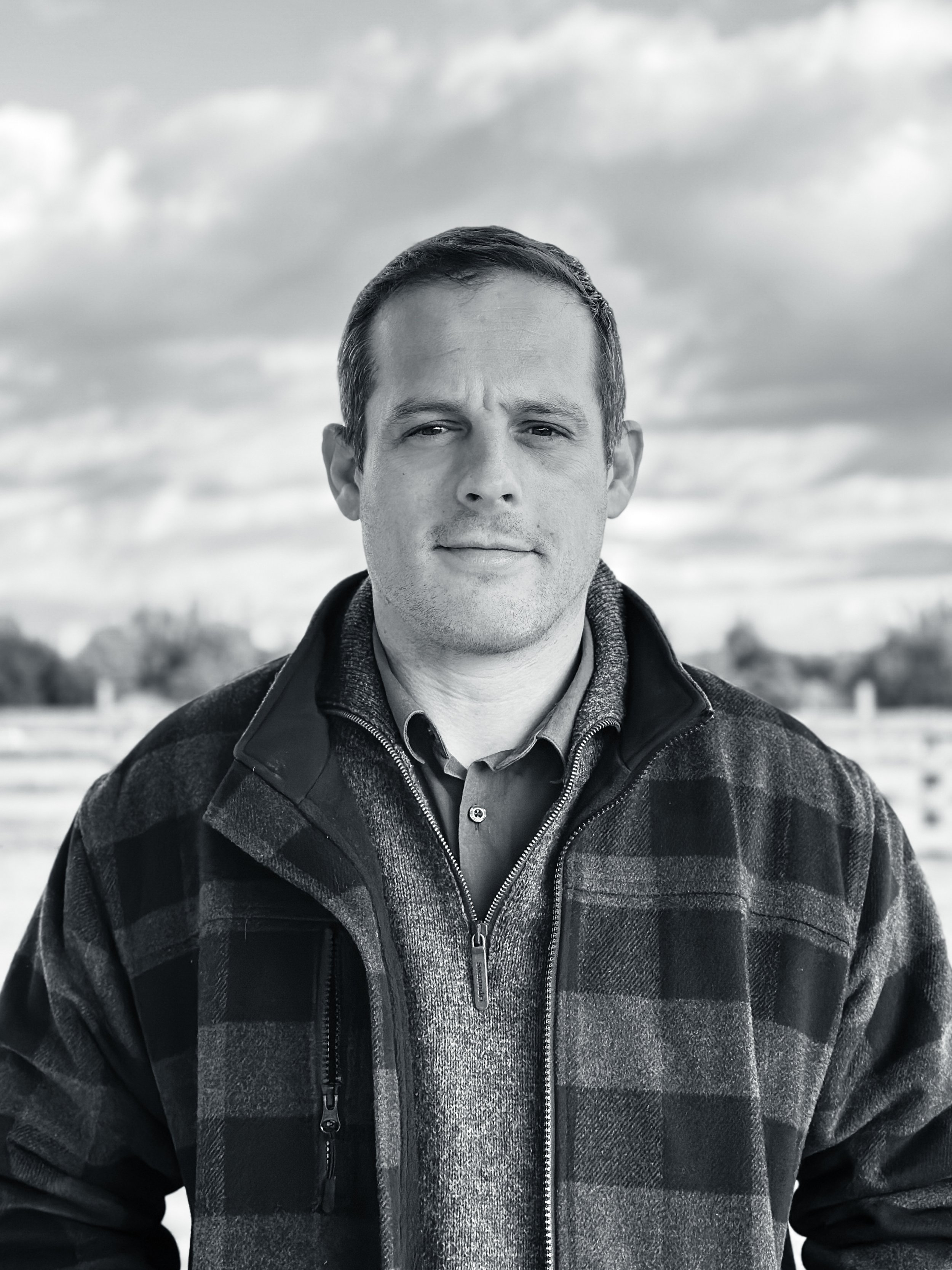 Jacques Jacobs - A black and white photo of a man outdoors, wearing a plaid jacket with a zipper, over a collared shirt, with a cloudy sky in the background.