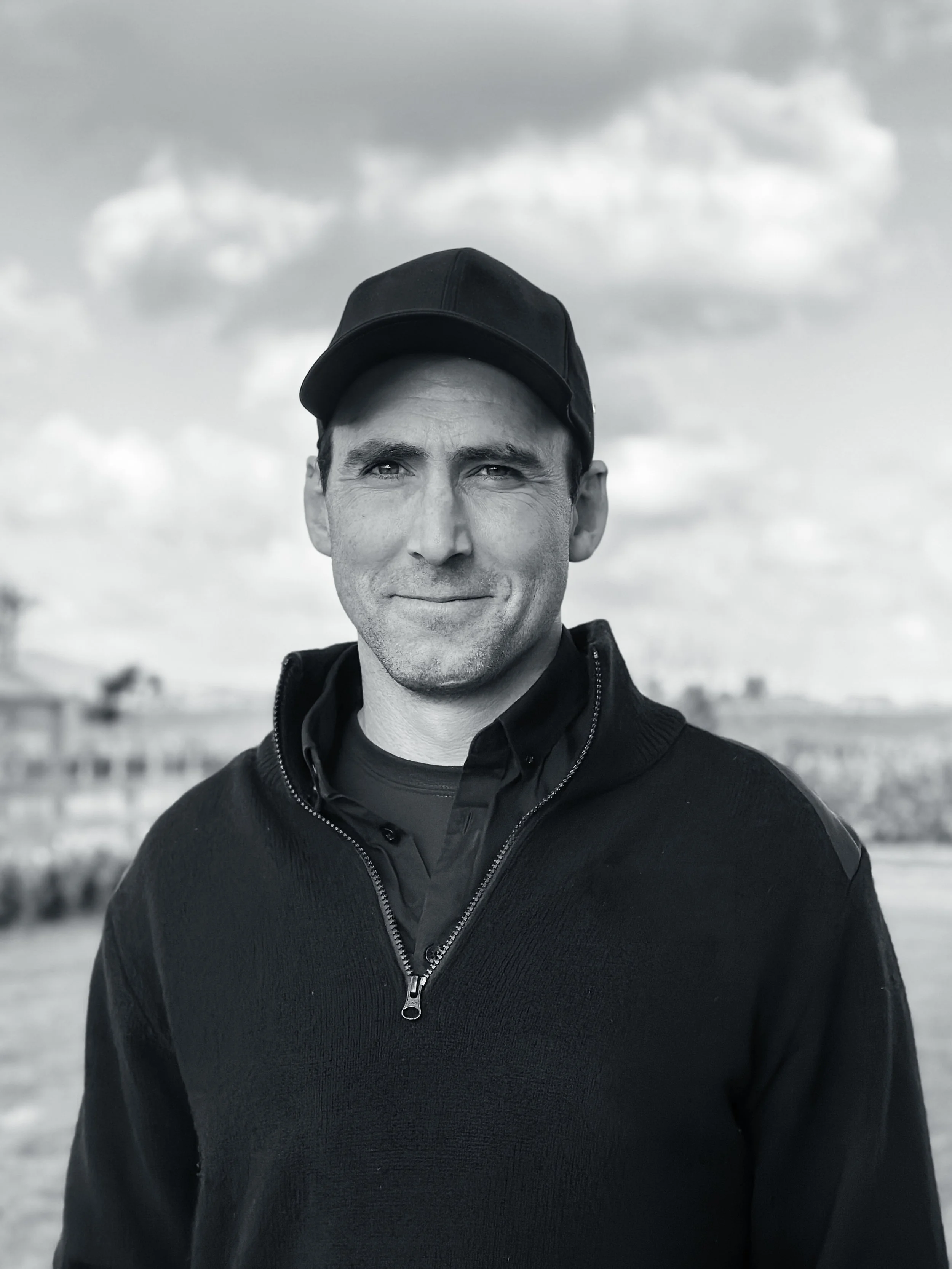 Sam Johnston - A man wearing a black cap and black jacket standing outdoors with cloudy sky in the background.