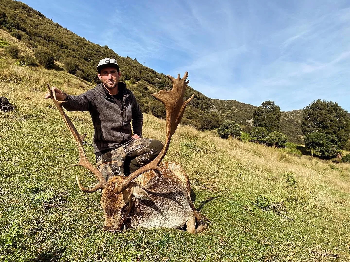 A man kneeling next to a large deer with antlers on a grassy hillside with trees and hills in the background.