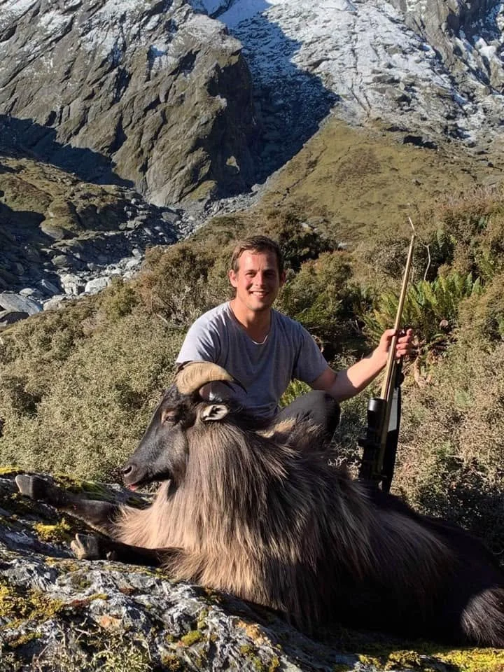 A man in a gray t-shirt kneeling with a rifle next to a large, dead mountain goat with curved horns in a mountainous landscape with green shrubbery and rocky terrain.