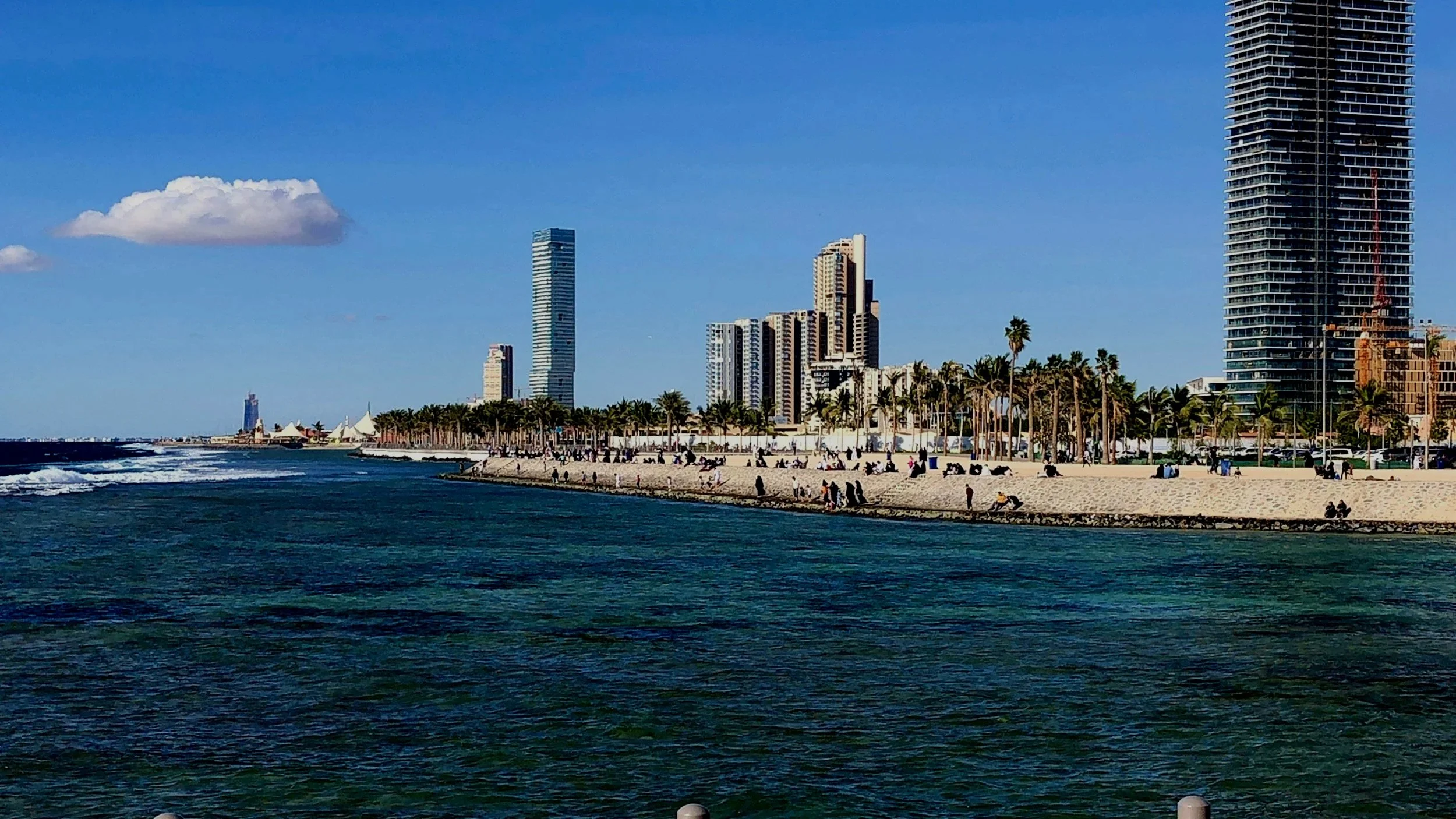 City skyline with tall modern buildings along a beach, with palm trees and people walking and relaxing on the sandy shore, under a blue sky with a few clouds.