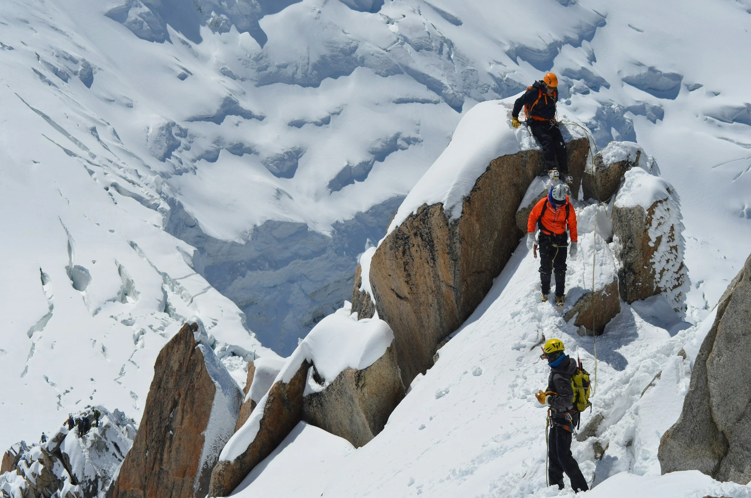 Mountaineers climbing snowy, icy mountain terrain with rocks, wearing helmets and harnesses, using ropes for safety.