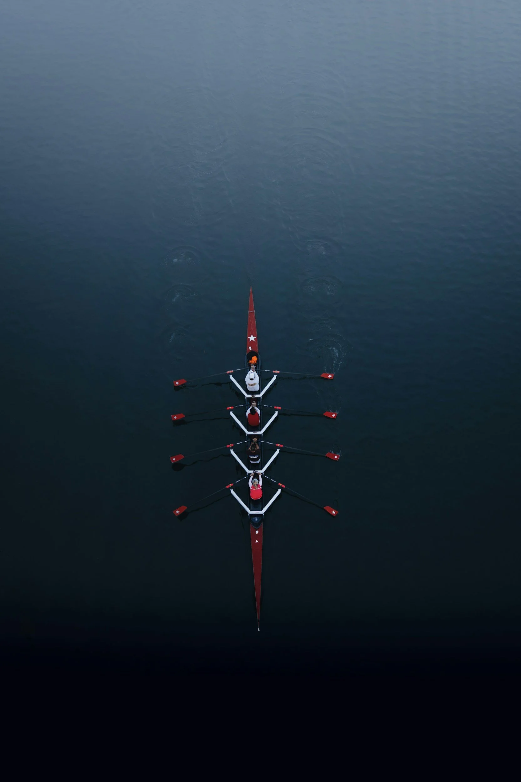 An aerial view of a rowing shell racing on open water with six rowers and a coxswain, with the water showing small ripples and occasional circles.