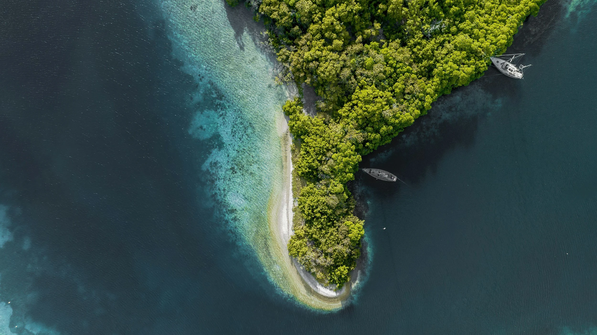 Aerial view of a small, forested island with clear blue water surrounding it, and two boats docked near the shoreline.