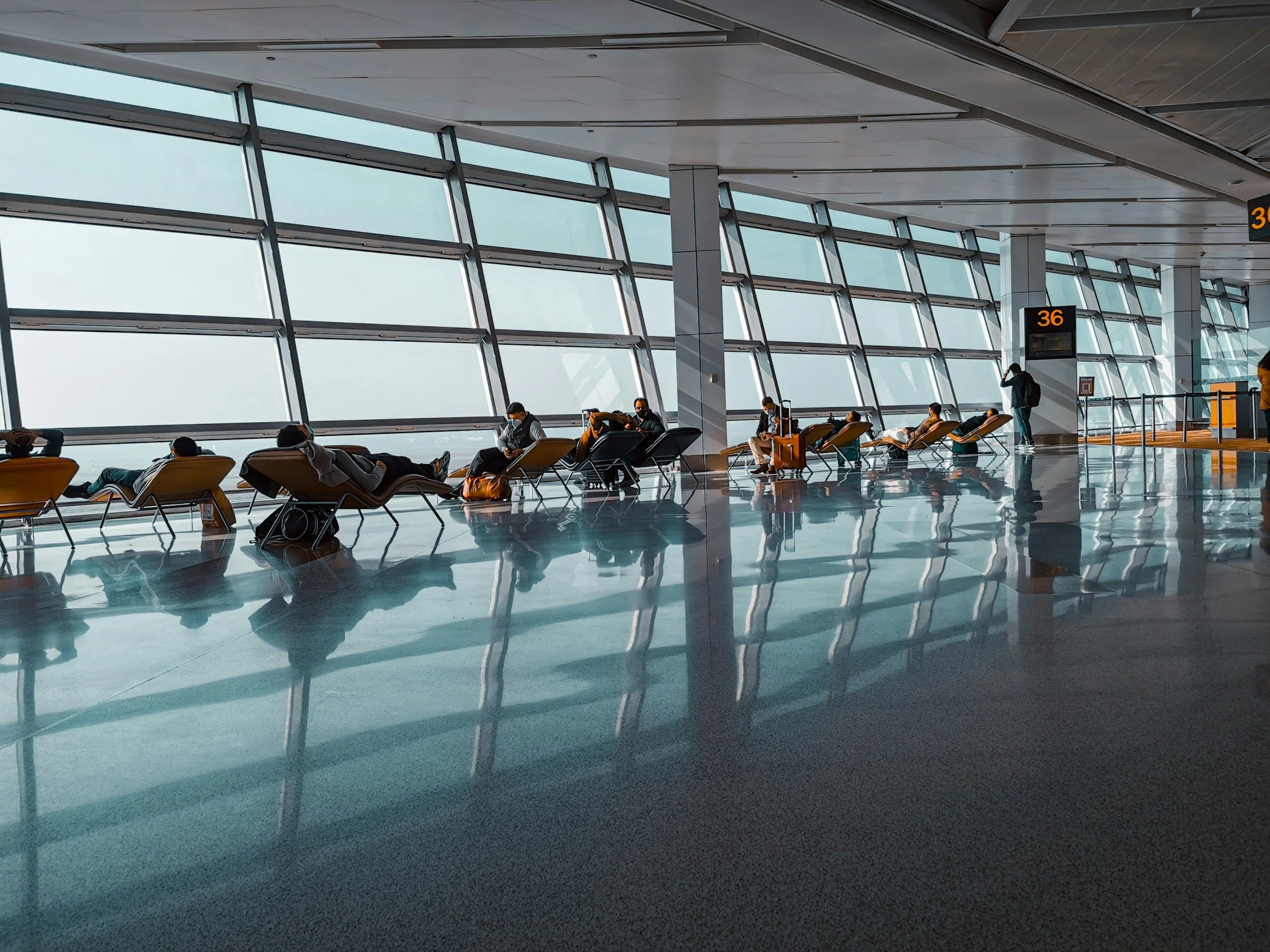 Interior of an airport terminal with large windows, passengers seated on chairs, some resting or using their phones, and digital screens displaying gate numbers.