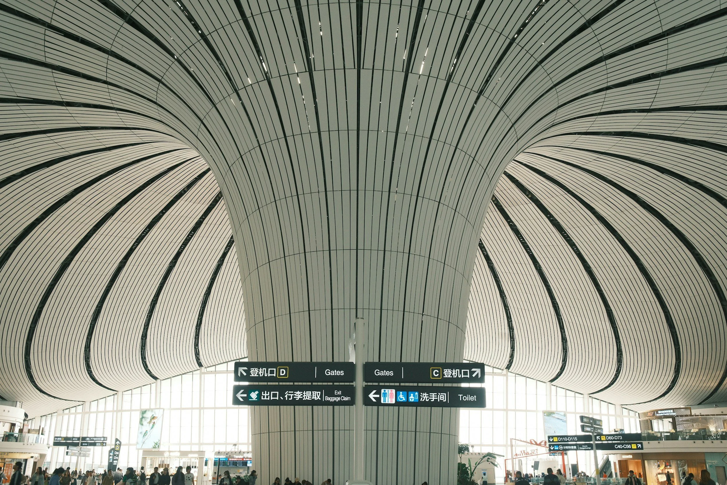 Interior view of a modern airport terminal with high arched ceilings, large windows, and overhead directional signs for gates, baggage claim, exits, and toilets. There are people walking and standing around.