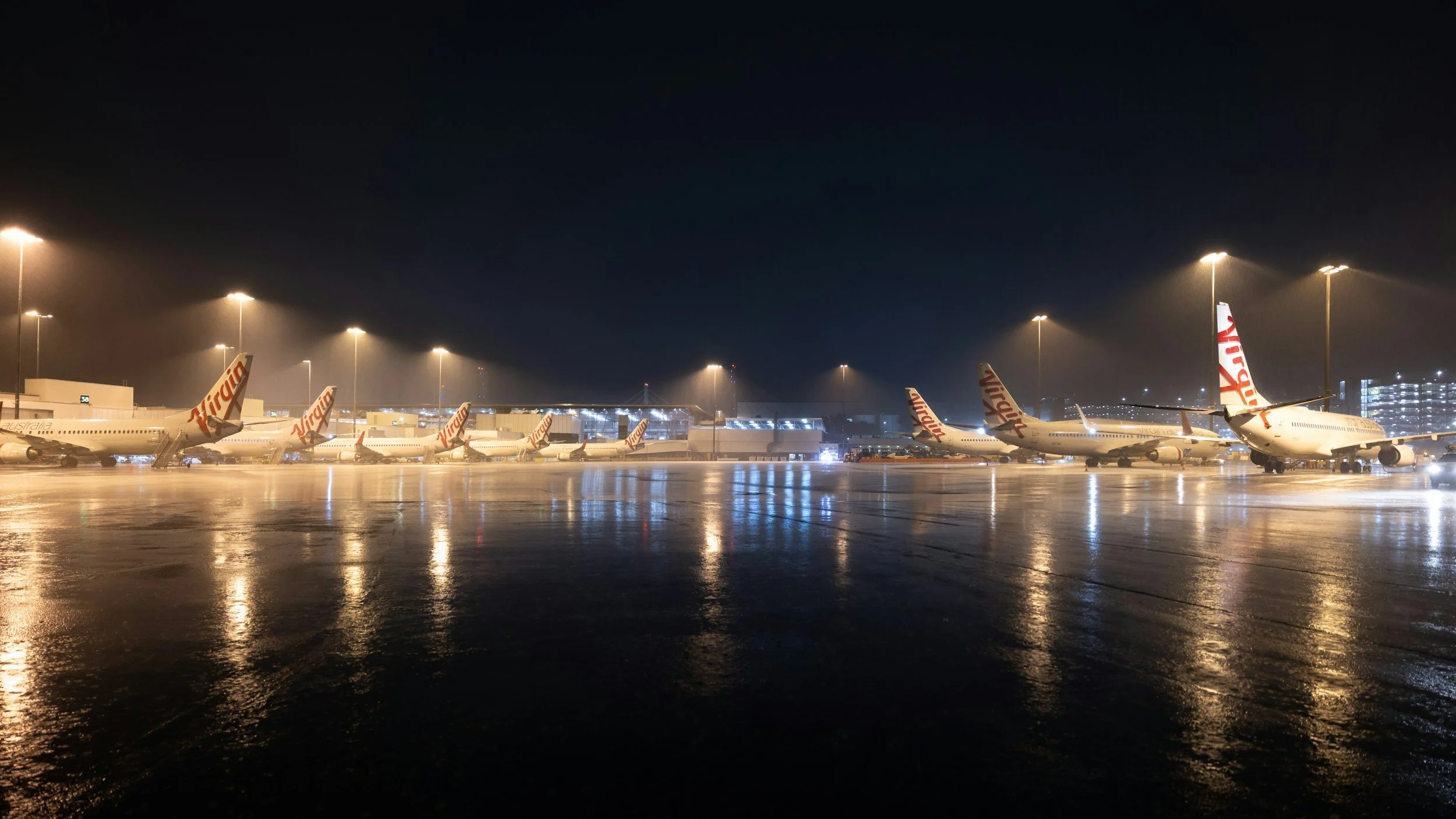 Multiple Virgin Australia airplanes parked on an airport tarmac at night, with their tails facing the camera and the airport illuminated by bright lights.