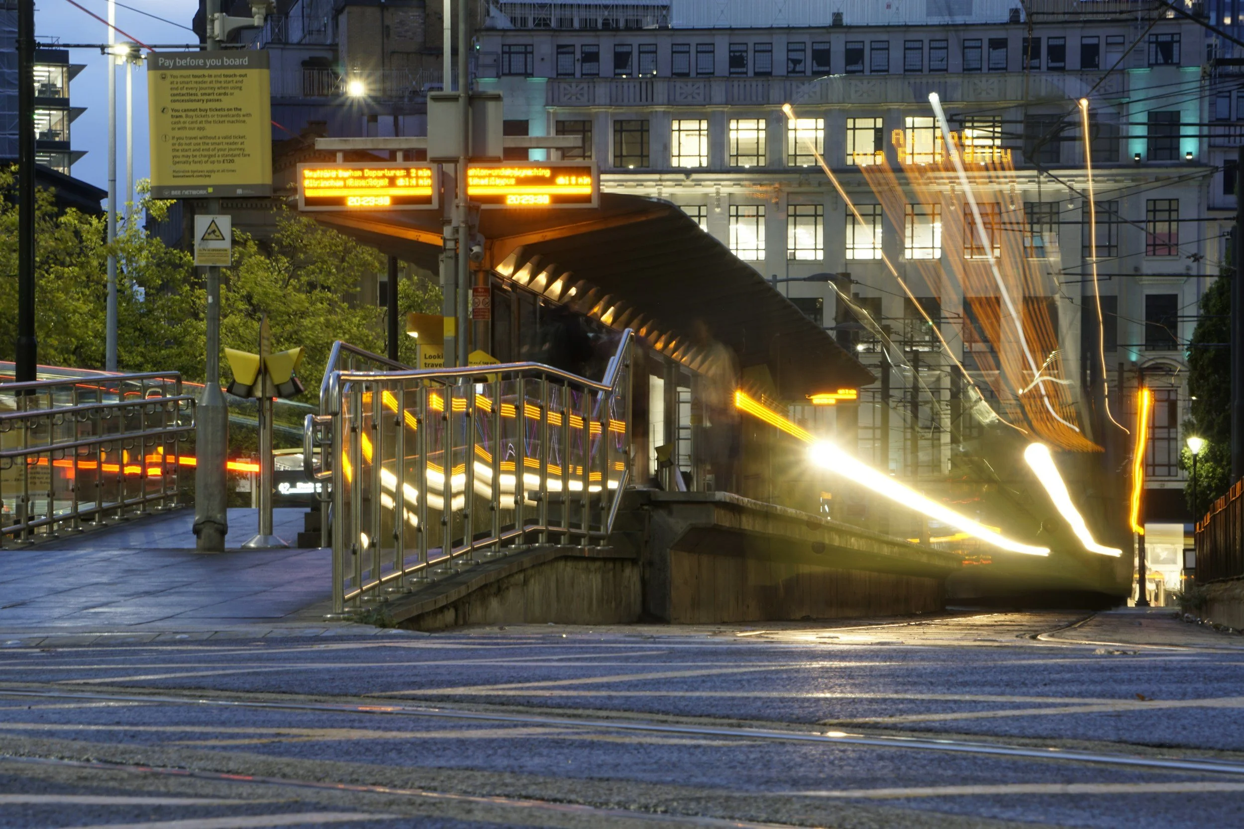 Urban scene at dusk with a train station entrance, illuminated signs, and motion-blurred train lights in the background.