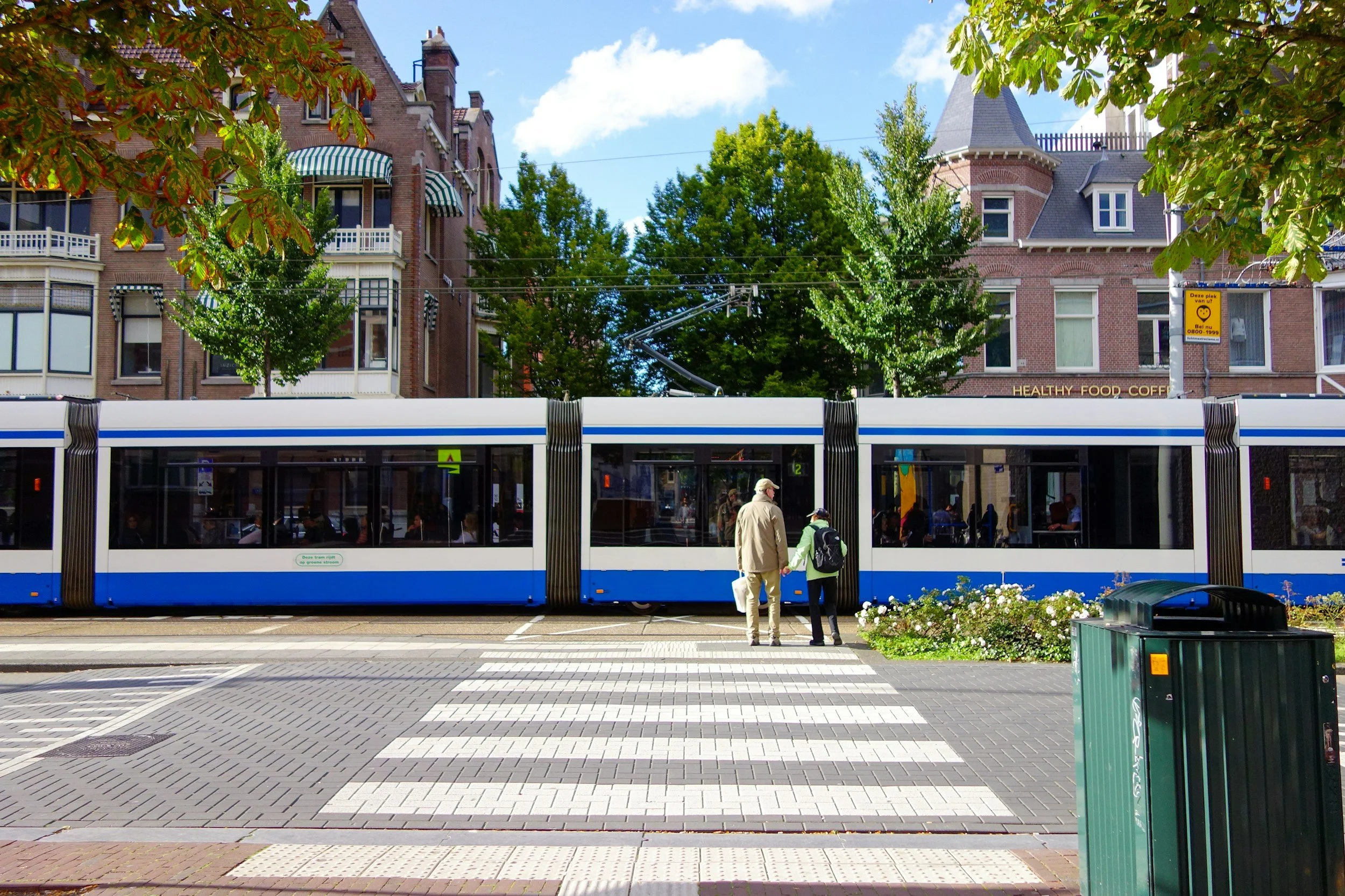 A tram stops at a street in a city, with two people waiting in front of it, surrounded by trees and historical brick buildings with turrets and striped awnings, under a partly cloudy sky.
