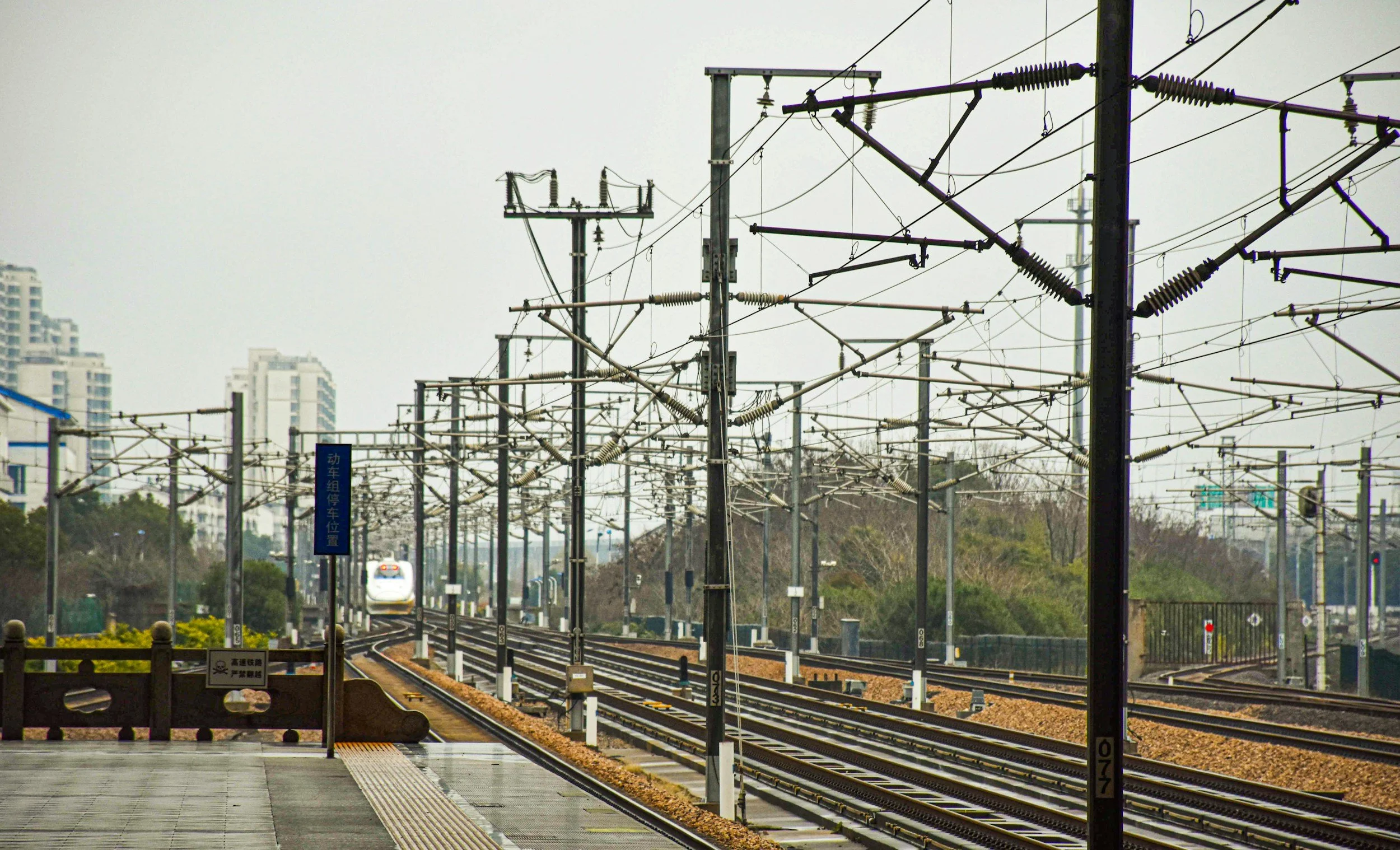 Multiple railway tracks with overhead electrical wires and signal signs, city buildings in the background, and a train approaching in the distance.