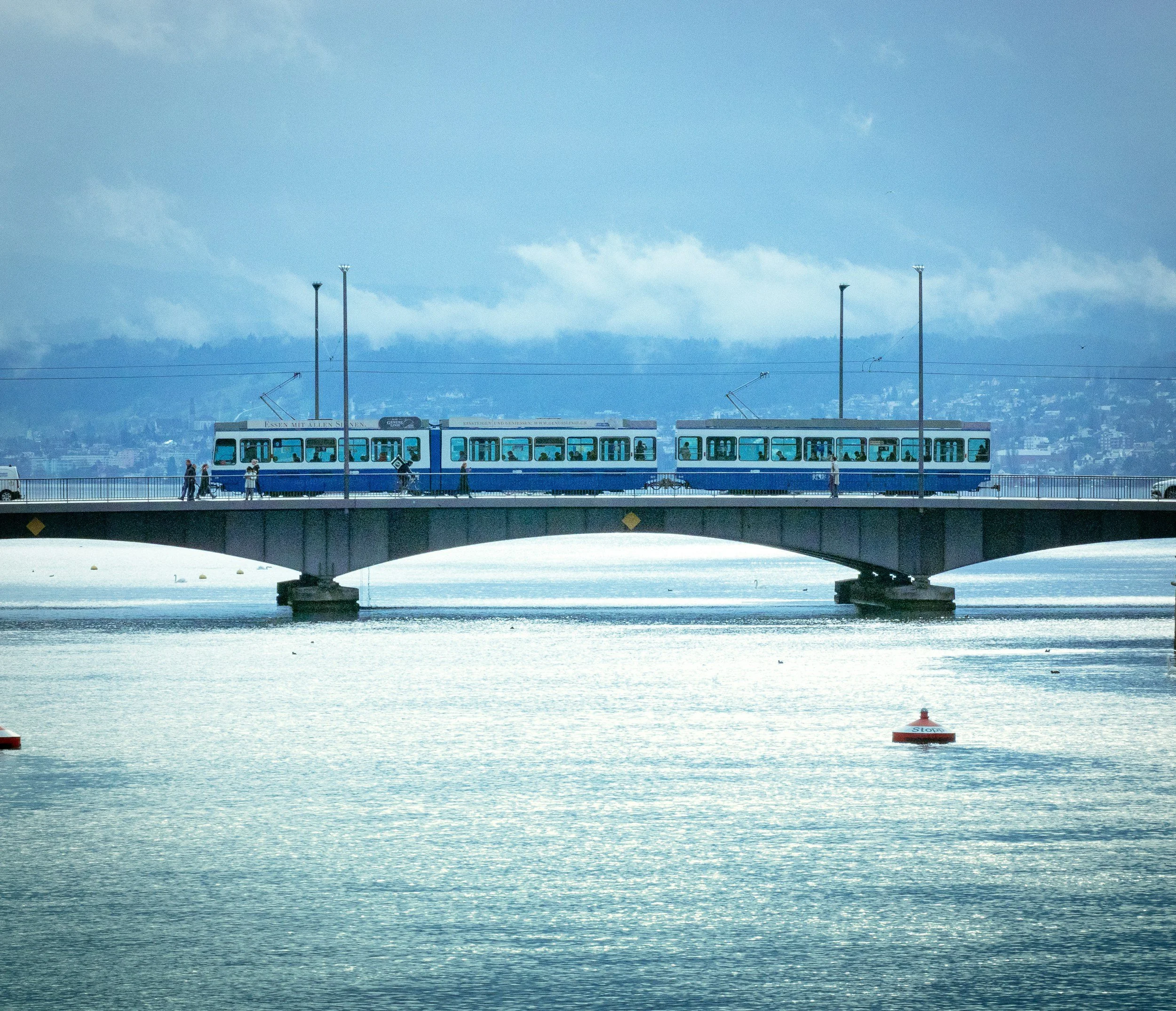 A blue and white tram on a bridge over a river with pedestrians walking along the bridge, under a blue sky with clouds and mountains in the background.