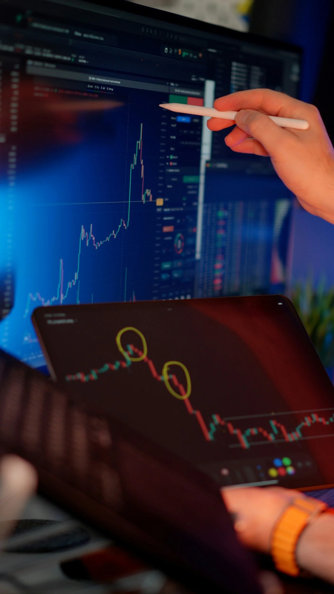 Person pointing at a computer screen with financial chart, second person holding a tablet with stock market charts, tracking financial data and stock performance.