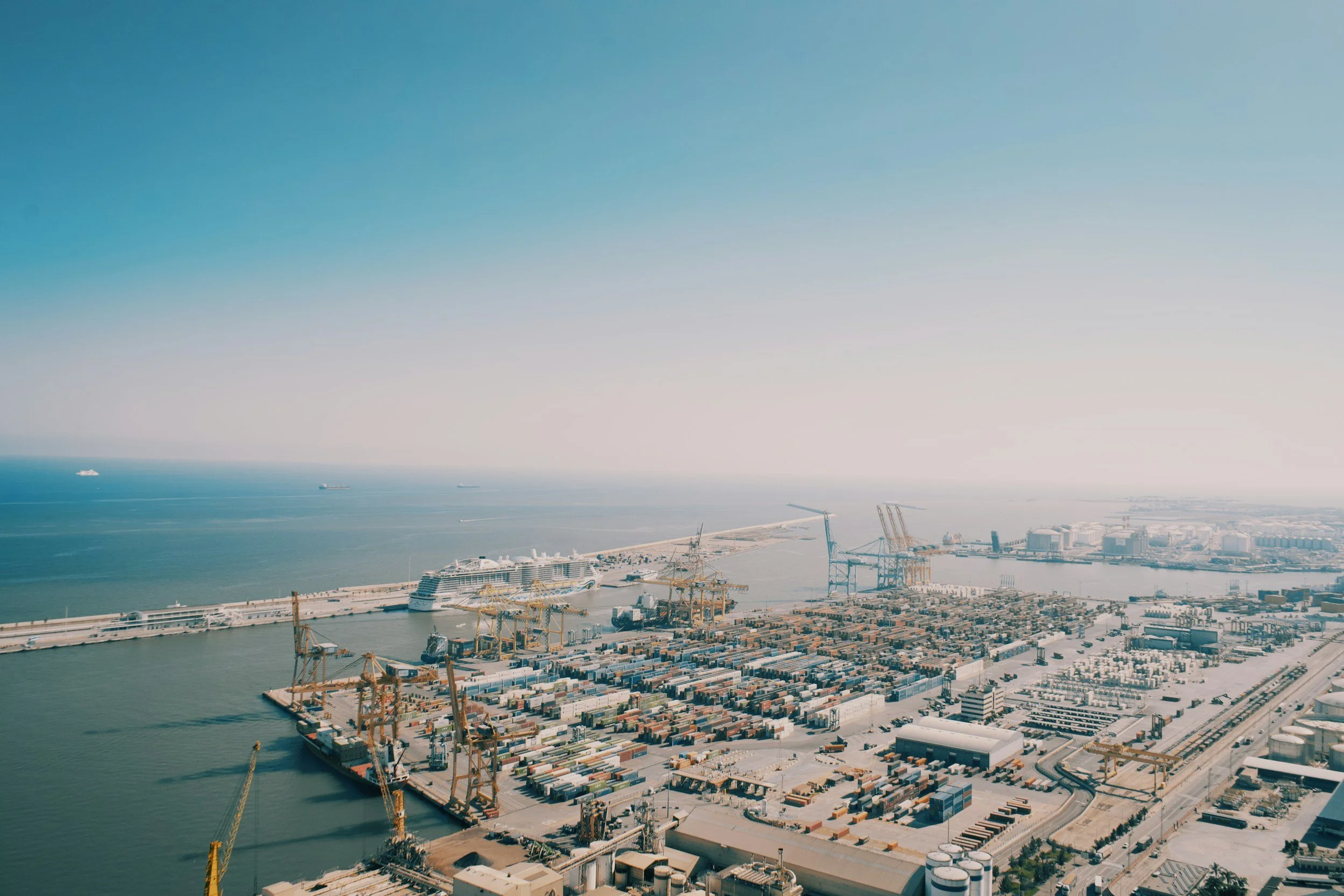 Aerial view of a port with cargo ships, numerous containers, cranes, and industrial buildings along the shoreline, with the ocean and a clear blue sky in the background.