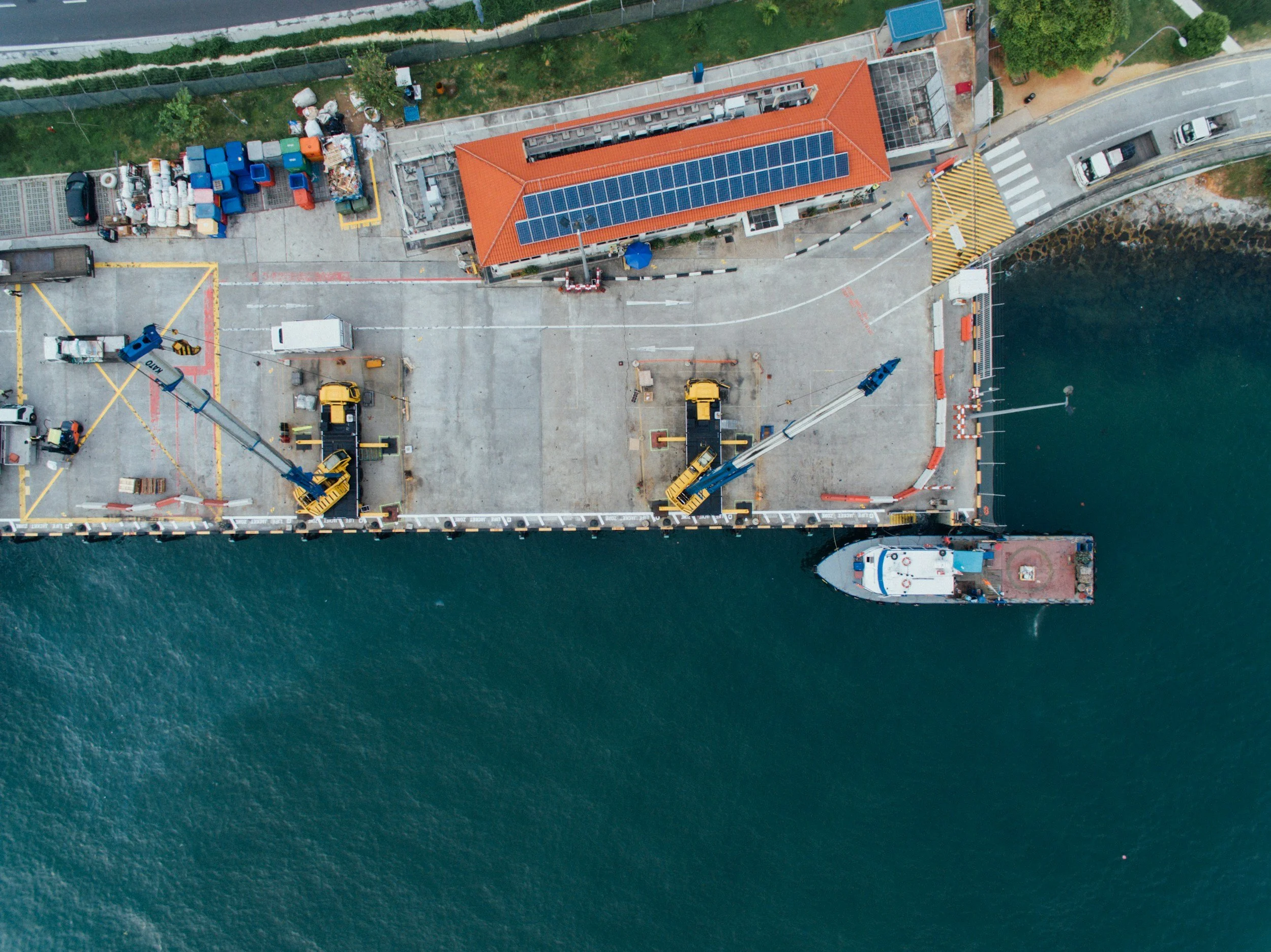 An aerial view of a harbor with a large ship docked near a concrete pier. The pier has construction equipment, cranes, and trucks, with a building that has solar panels on its orange-tiled roof. Adjacent to the harbor is a green area separated by a fence, and a street with parked cars and a pedestrian crossing.
