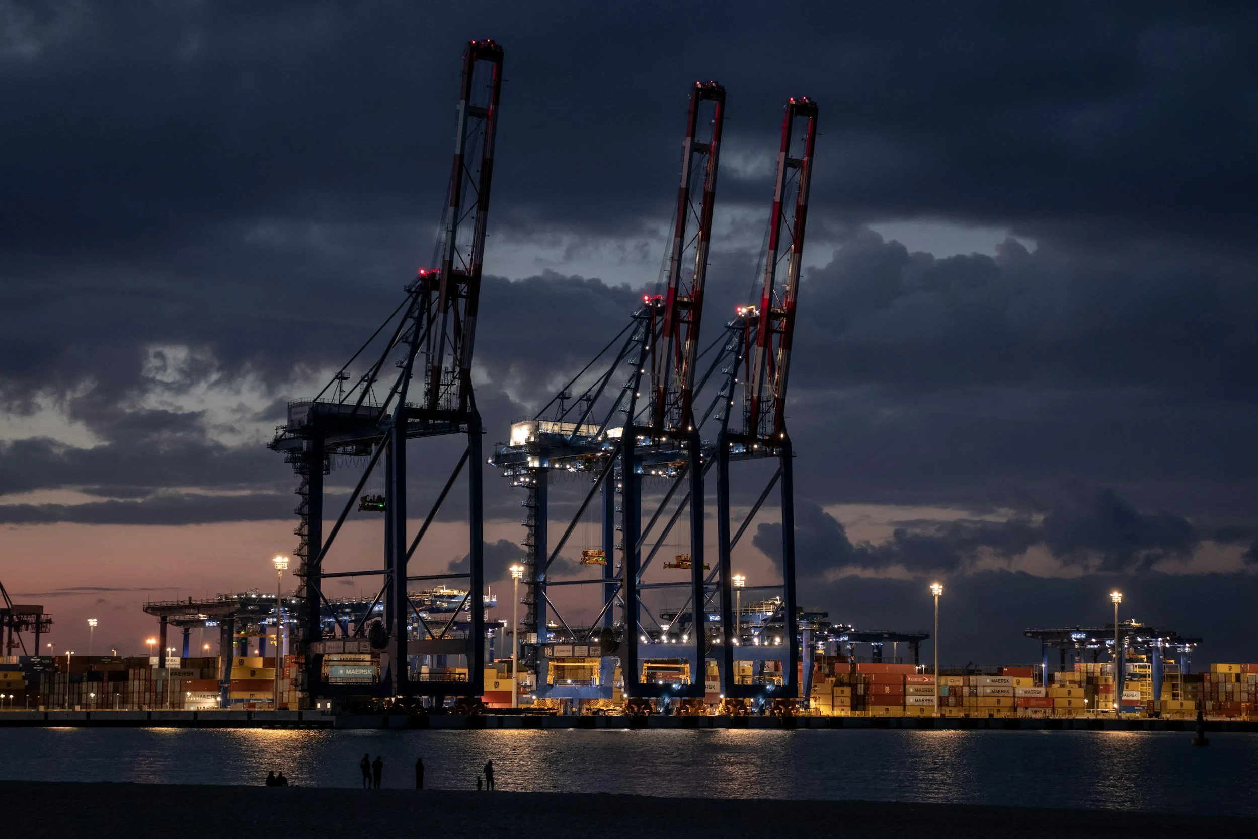 Container cranes at a shipping port during dusk, with stacked containers below and a dark cloudy sky overhead.