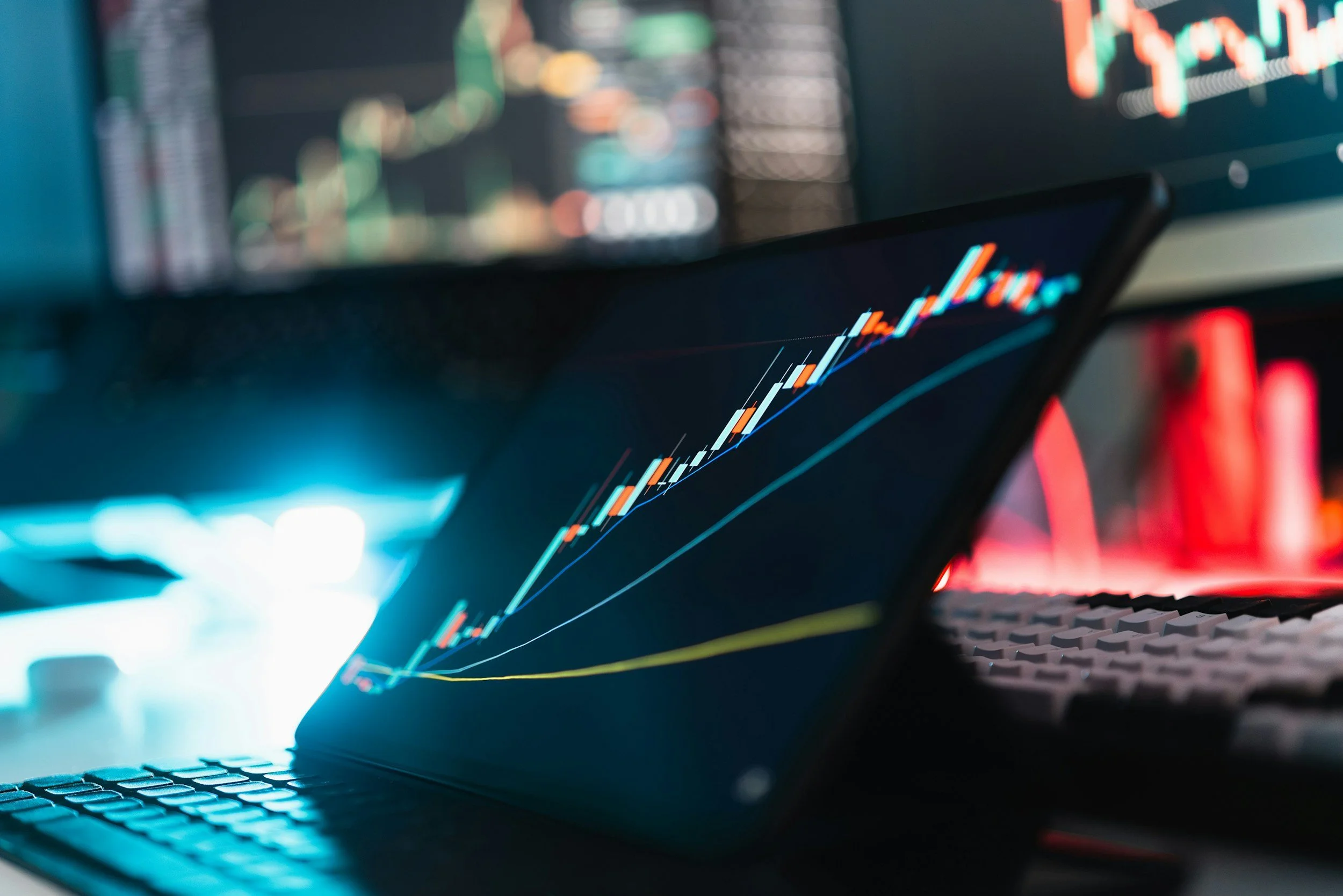 Close-up of a computer screen displaying colorful stock market or financial trading charts with candlestick patterns and trend lines, with a blurred background of a keyboard and a desk setup.