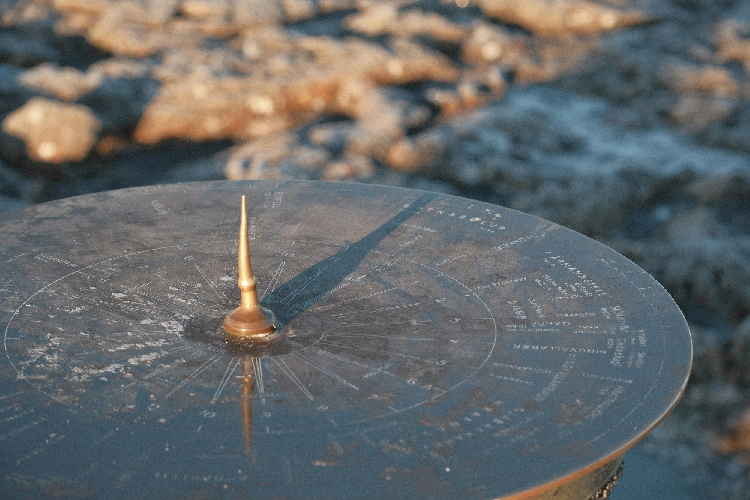 Close-up of a navigational compass on a boat, with rocky shoreline water blurred in the background.