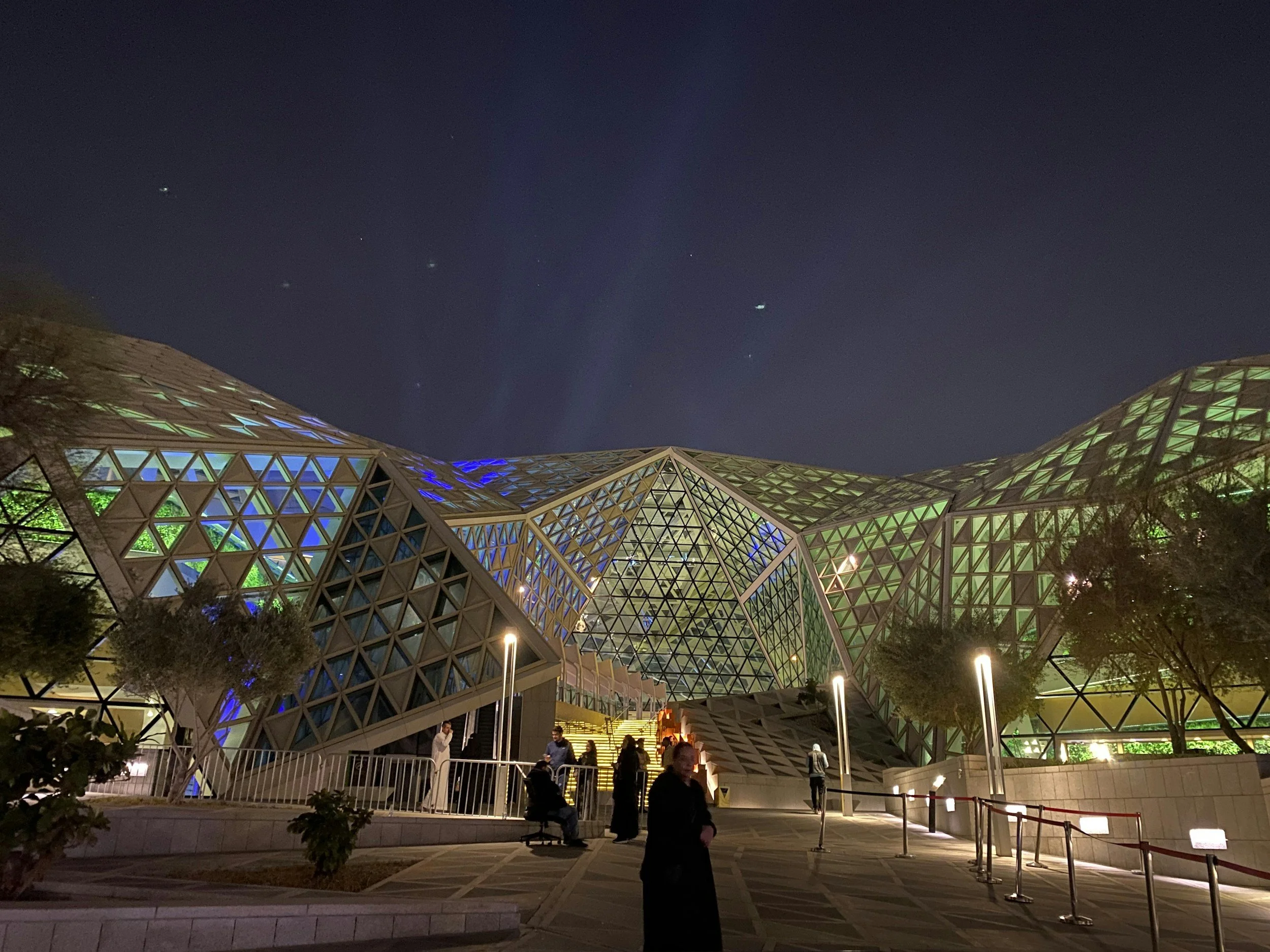 Night view of modern architectural buildings with glass and geometric designs, illuminated with colorful lights, with people sitting and walking outside under a starry sky.