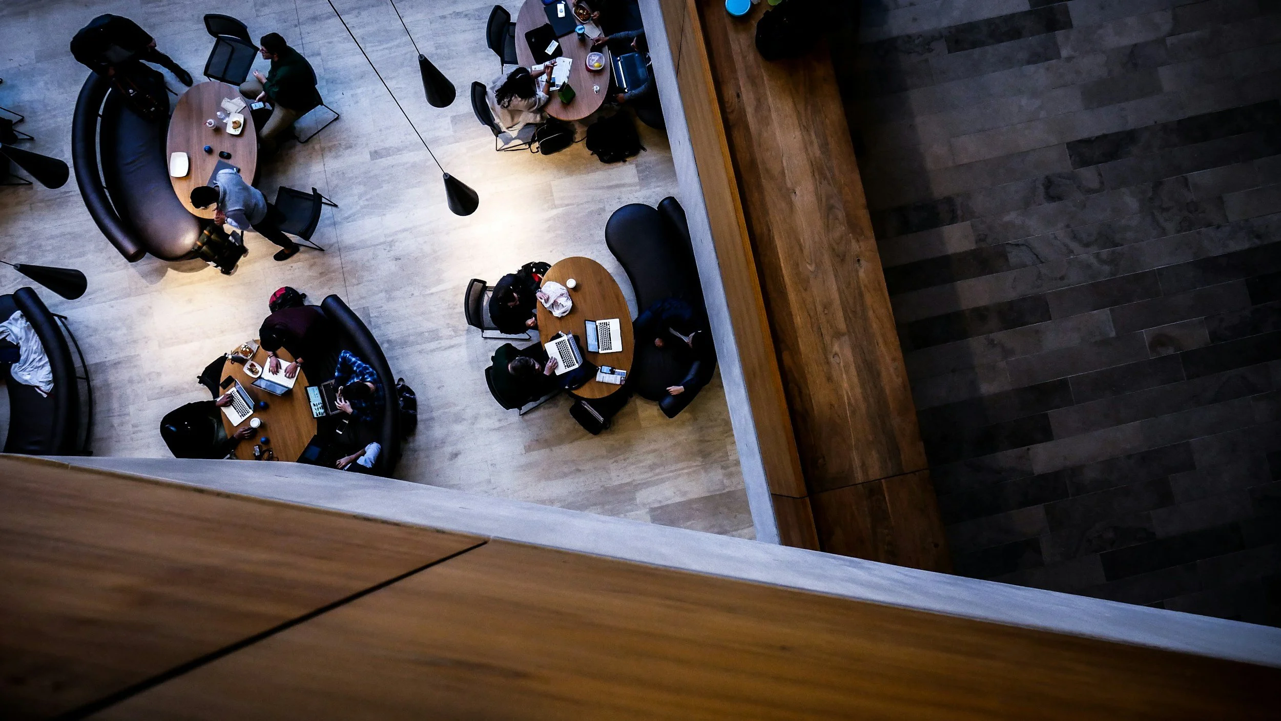 Overhead view of a modern, multi-person workspace with some people working on laptops and others drinking coffee, surrounded by tables and chairs in a spacious, well-lit office area.