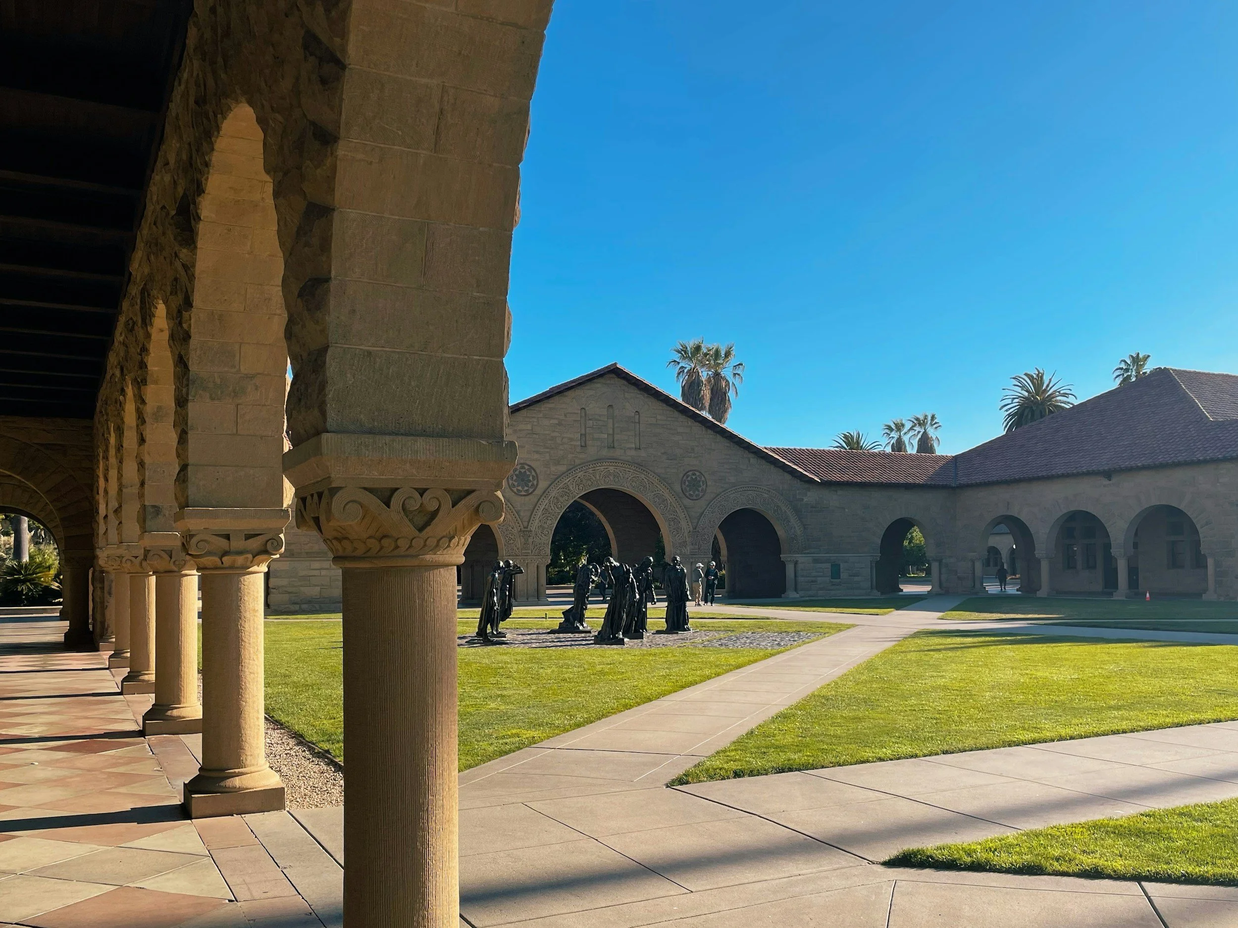 A courtyard with stone columns on the left, a building with decorative arched windows and palm trees in the background, and a row of sculptures of standing figures in the center.