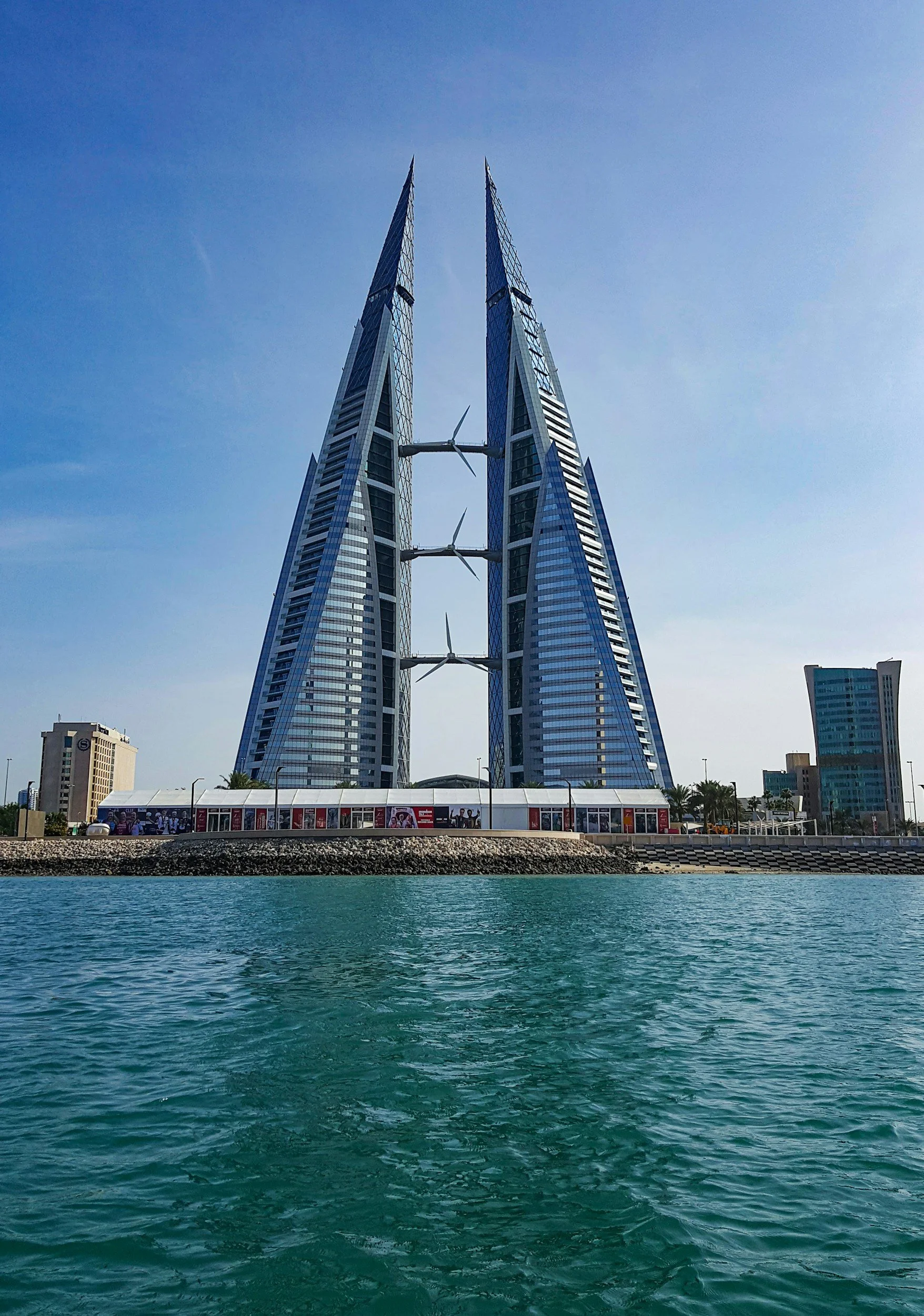 The image features the silhouetted, modern architecture of the famous Bahrain World Trade Center, consisting of two twin towers with wind turbines connecting them, located over a body of water on a clear, sunny day.