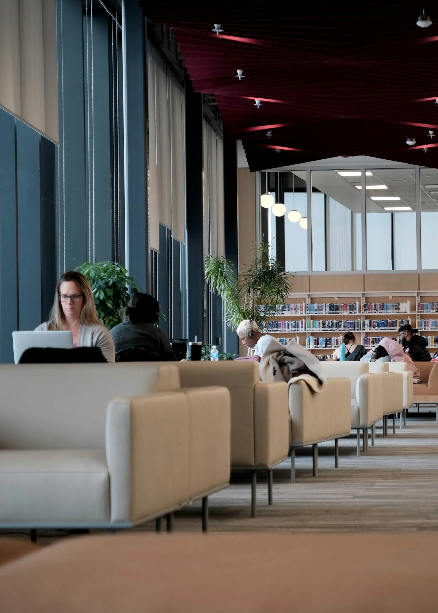 Interior of a modern library or study area with large windows, beige seating, and people using laptops and listening to headphones.