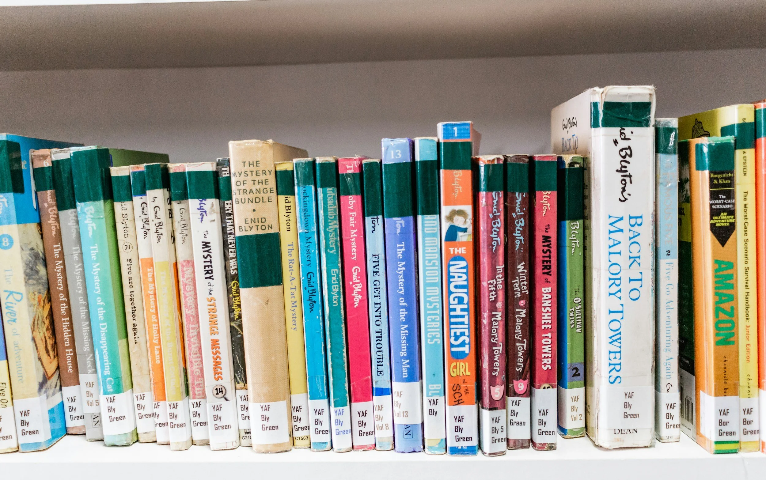 A shelf filled with colorful young adult fiction books, with titles including 'The Mystery of the Strange Bundle' and 'Back to Malory Towers'.