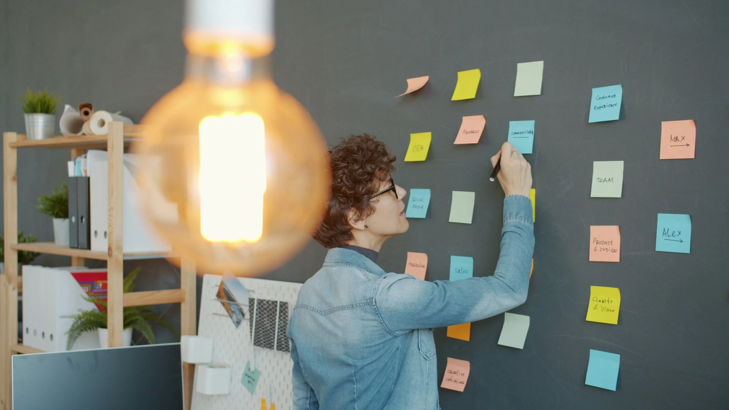 Woman with curly hair and glasses writing on sticky notes attached to a black wall in an office.