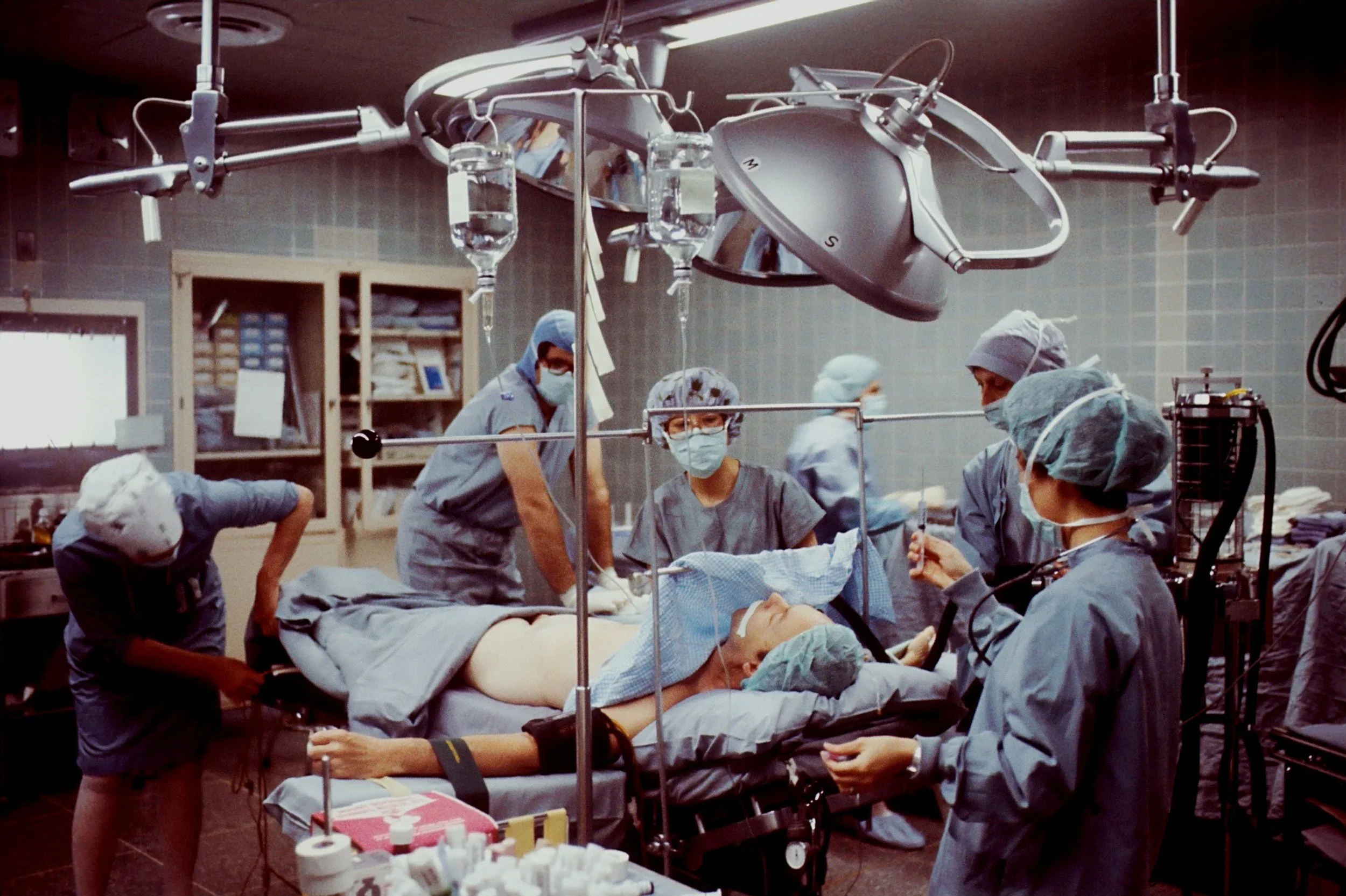 A team of surgeons and medical staff performing surgery in an operating room, with medical equipment and monitors around them.