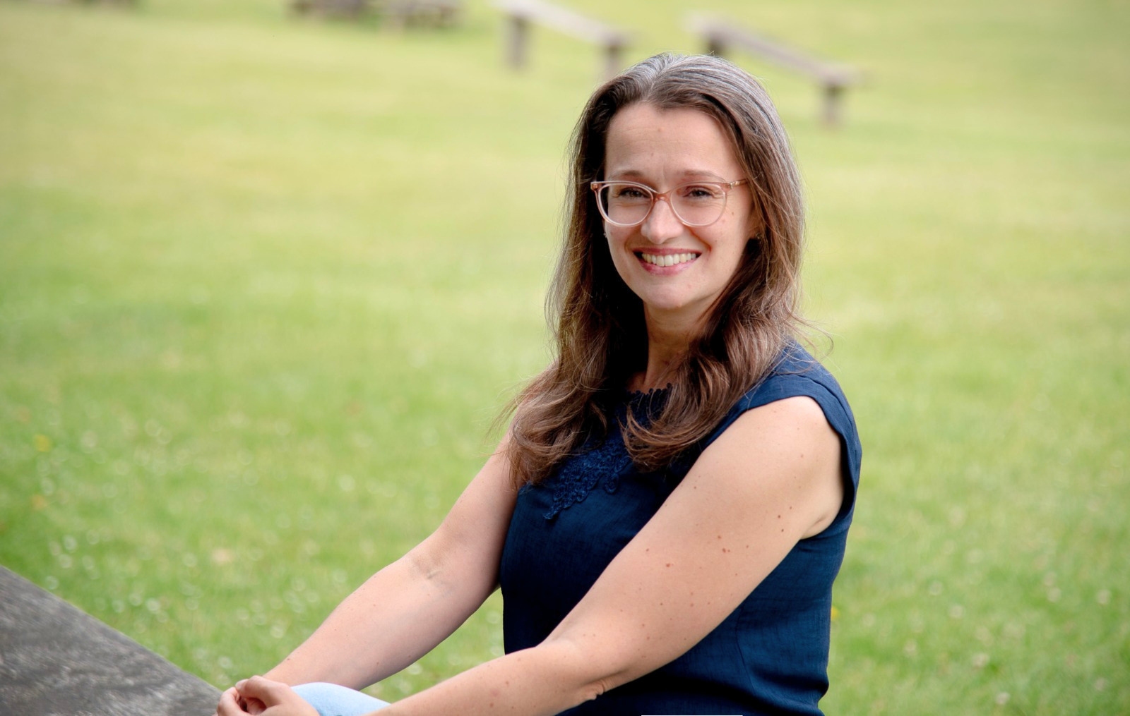 A woman with long brown hair, glasses, and a blue sleeveless top sitting outdoors on a grassy field with blurred benches in the background.