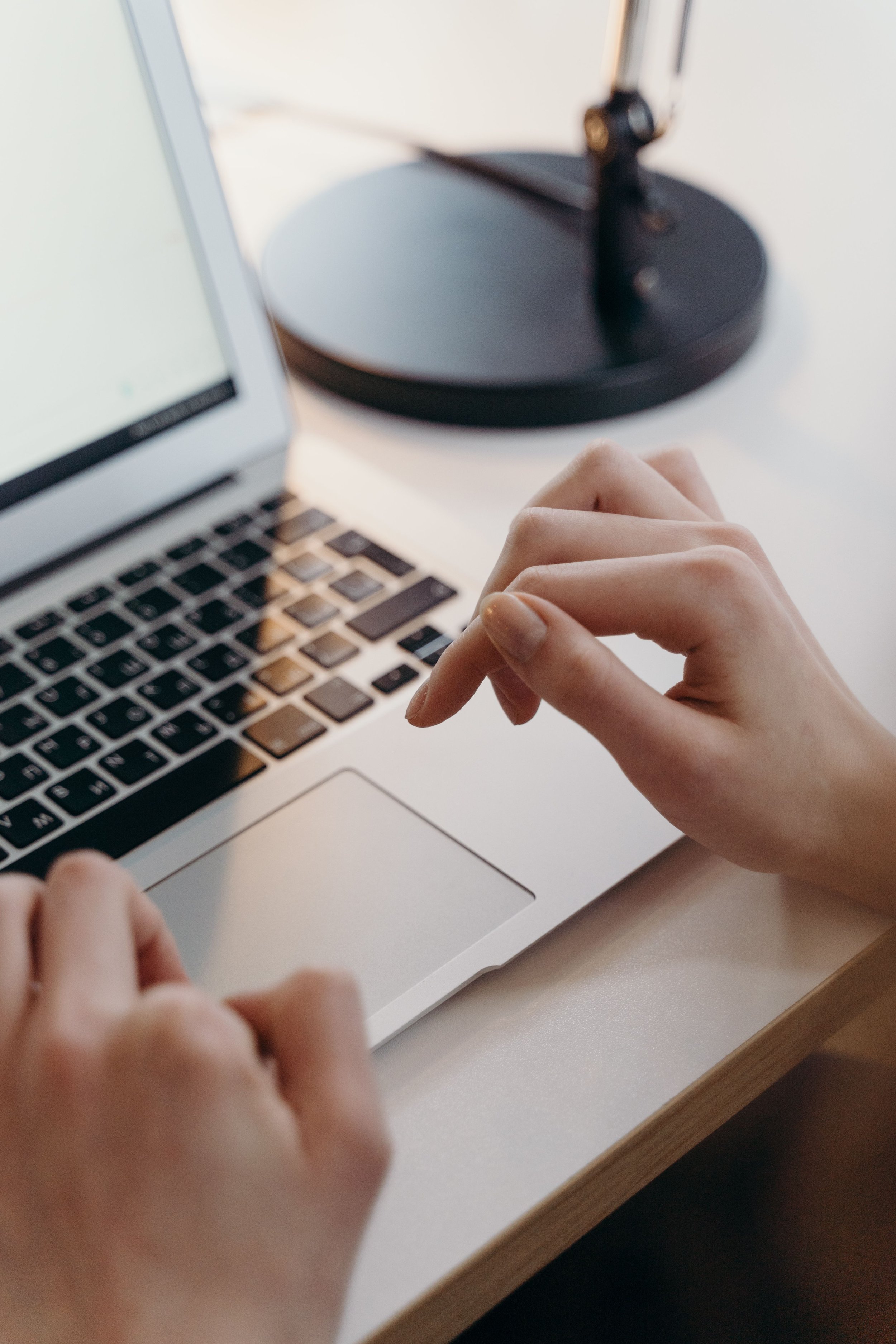 Close-up of hands typing on a MacBook keyboard with a computer monitor and a black desk lamp in the background.