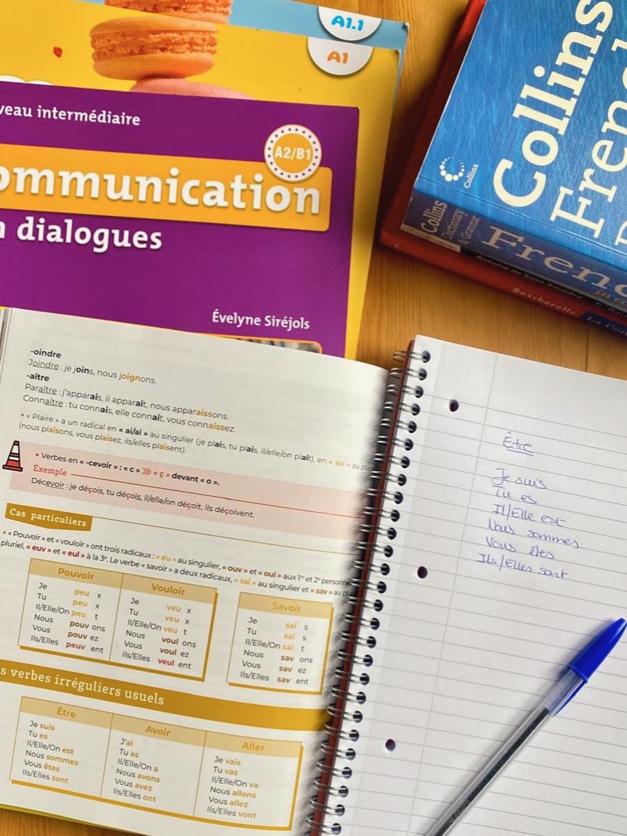 Open language learning French books and notes on a wooden table, including a purple book titled 'Communication in dialogues,' a blue book about 'Collins English - French,' and a notebook with handwritten French notes.