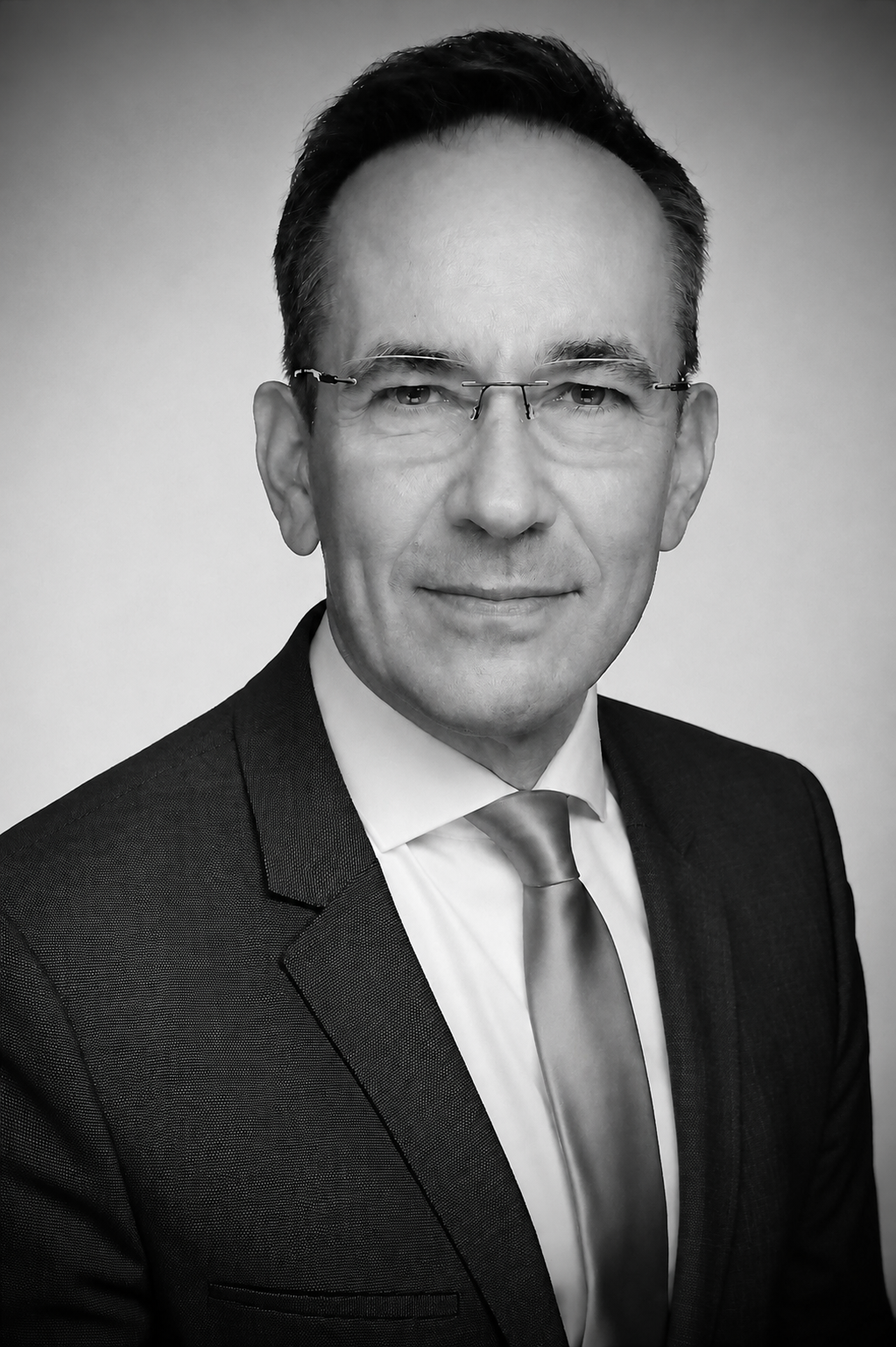 Schwarz-weiß Porträt eines Mannes im Anzug mit Krawatte und Brille, lächelnd, vor hellem Hintergrund.