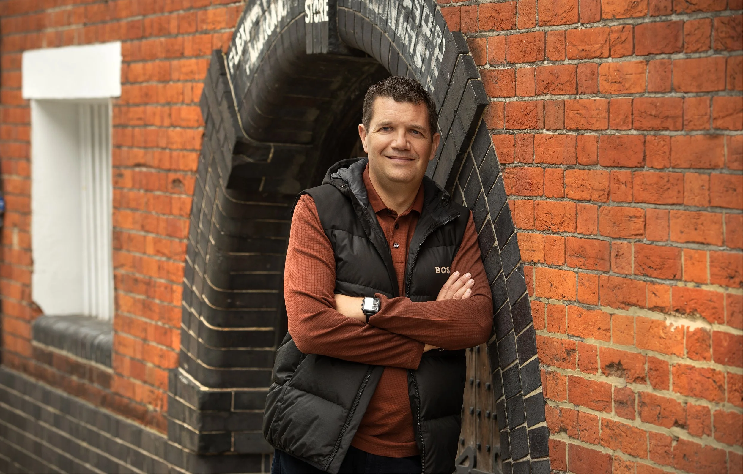 A man in a brown shirt and black vest crossing his arms, standing in front of a brick wall with an arched window.