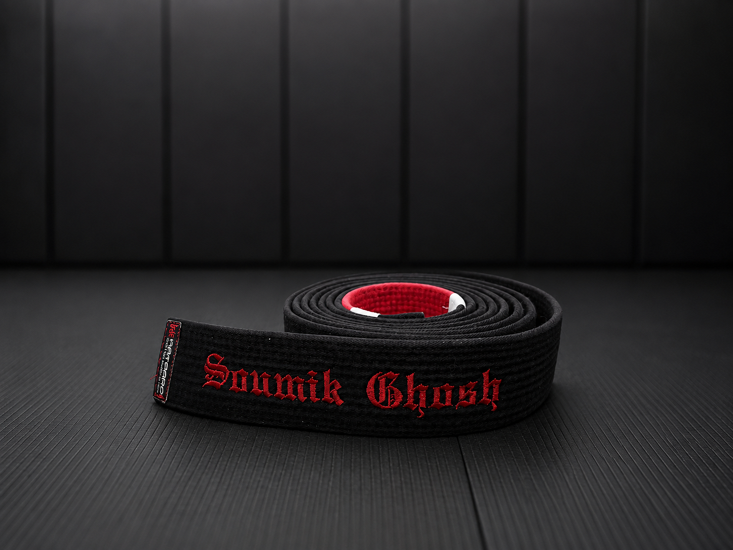 A black Brazilian Jiu-Jitsu belt with red embroidery reading 'Solounik Ghoash' laid on a black textured gym floor with a black padded wall in the background.