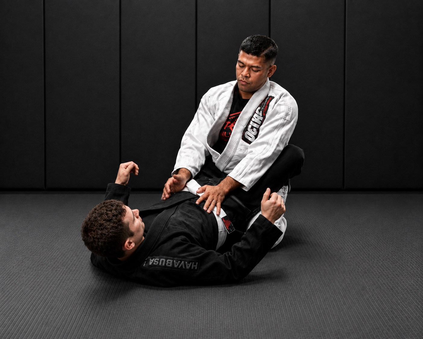 Two men practicing Brazilian Jiu-Jitsu, with one man in a white gi on top and the other man in a black gi on the bottom, inside a gym with black padded walls.