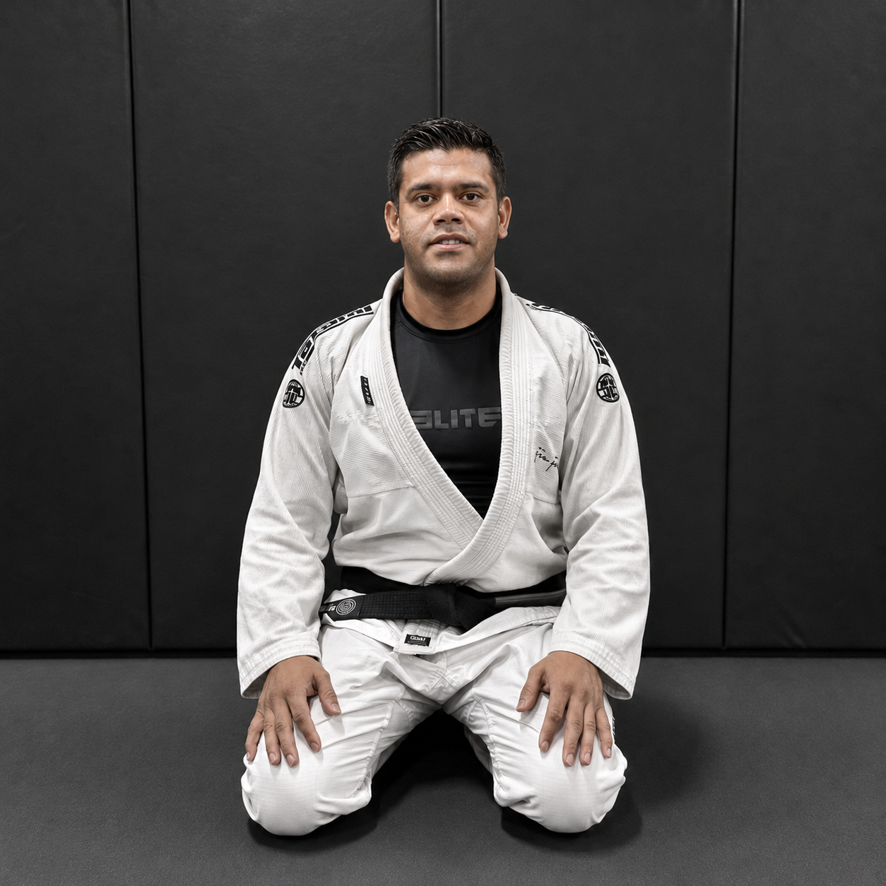 A man in a white Brazilian Jiu-Jitsu gi kneeling on a black mat against a black wall.