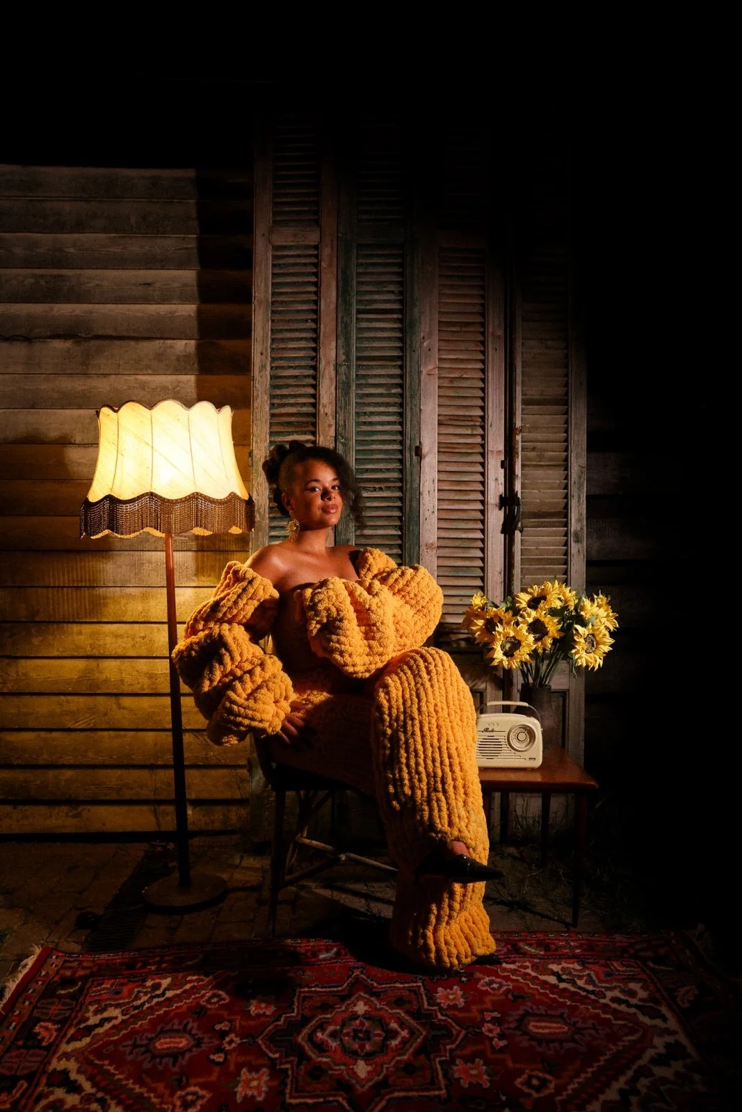 A woman dressed in a cozy, bright yellow, textured outfit sitting in a rustic room with wooden walls and shutters, next to a vintage lamp, a bouquet of sunflowers, a radio, and a patterned rug.