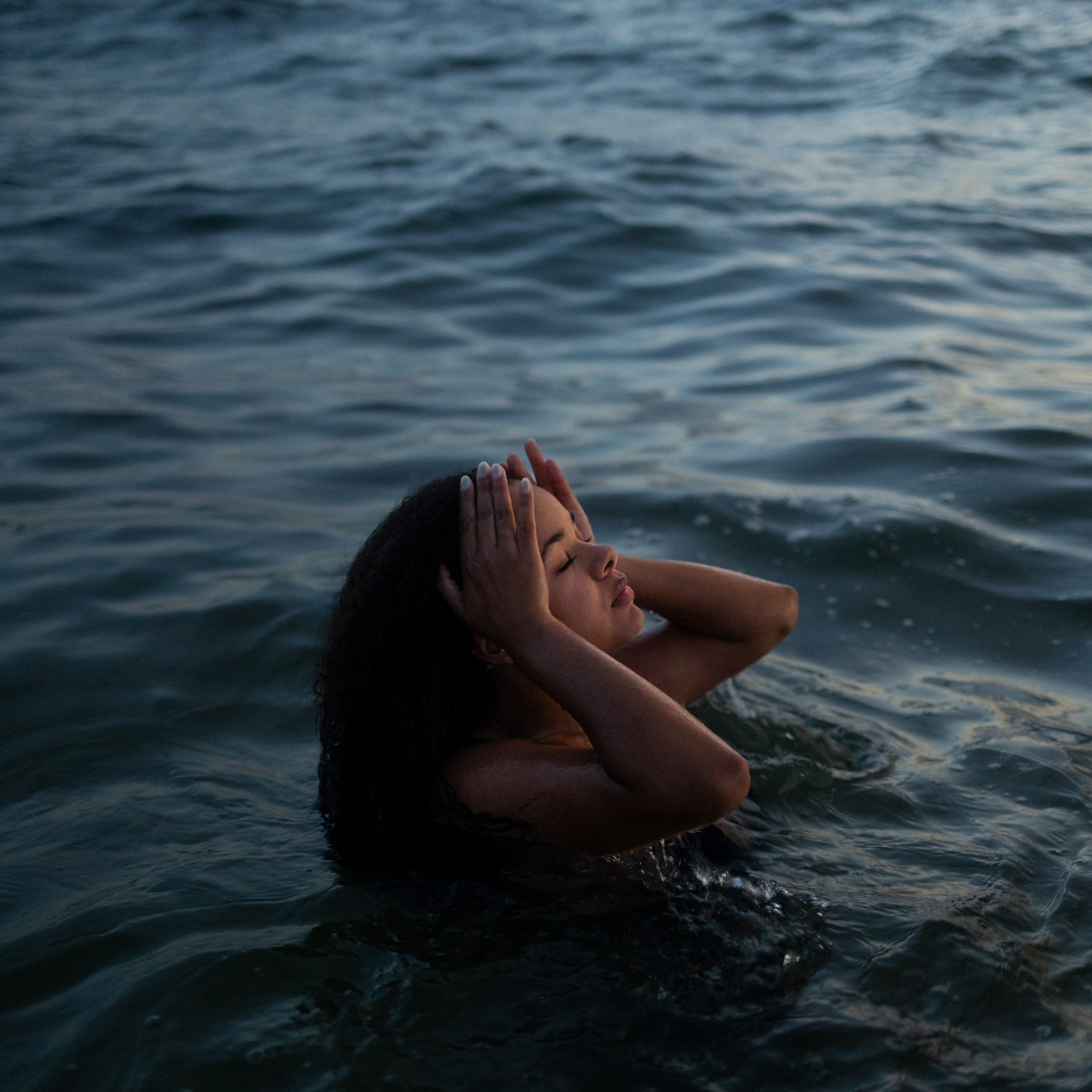 A person floating in a body of water with their eyes closed and hands on their head, surrounded by rippling water surfaces.