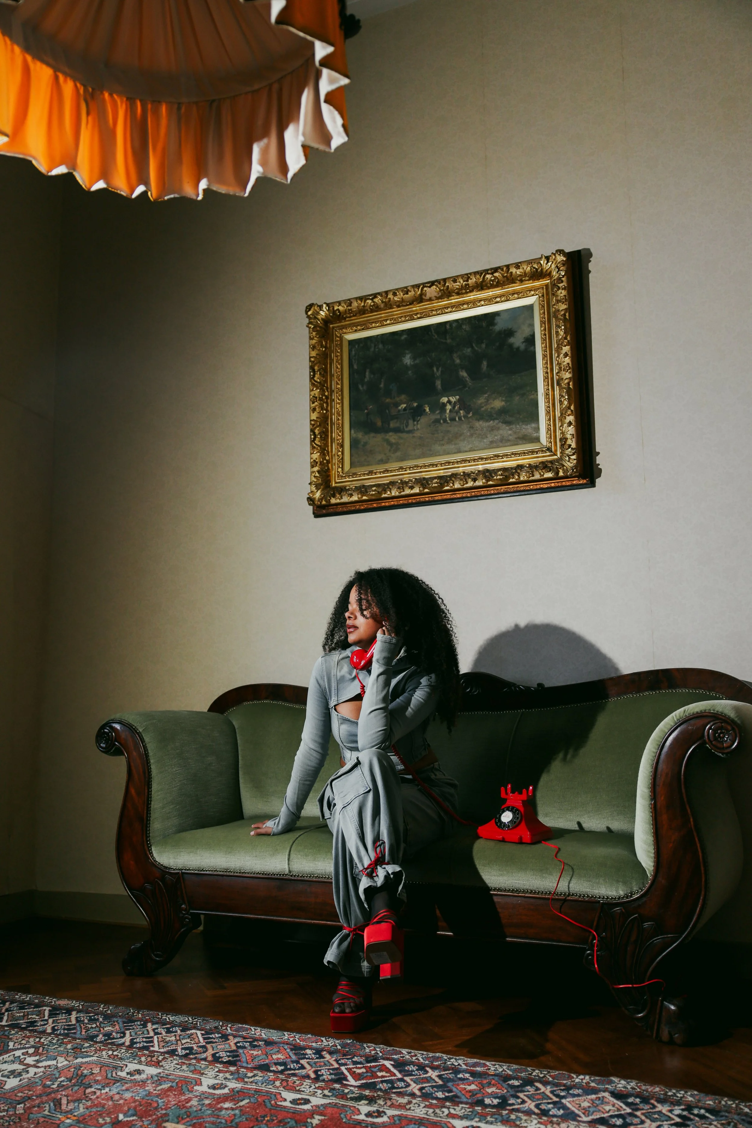 A woman with curly hair wearing gray clothes and red platform shoes sitting on a vintage green couch, holding a red corded vintage rotary phone. There is a painting of cows and a cart hanging on the wall behind her, and an orange lampshade hangs from the ceiling. Rugs are on the floor.