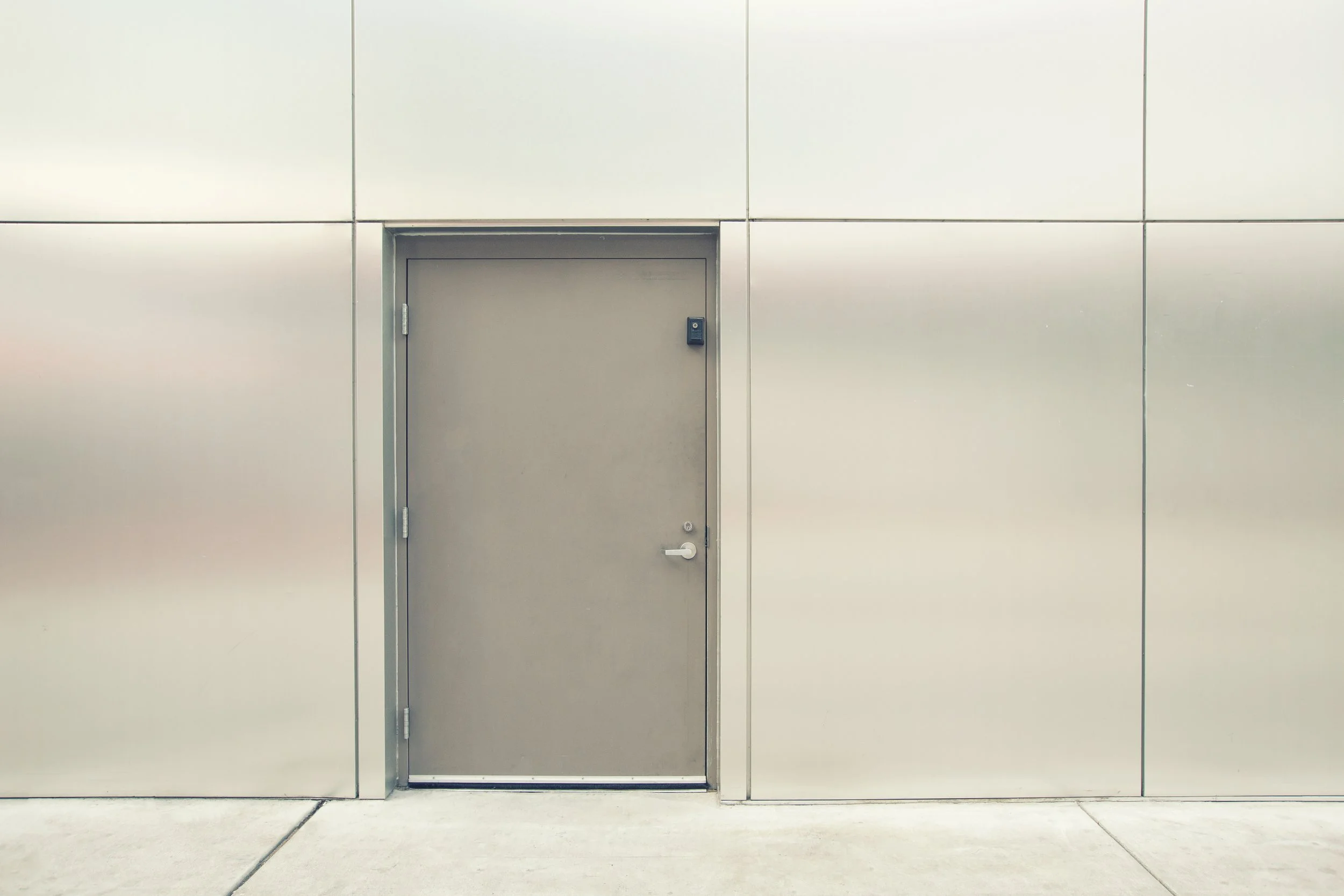 A plain gray personnel door with a lock and handle, set into a white wall with visible panel lines, against a concrete sidewalk.