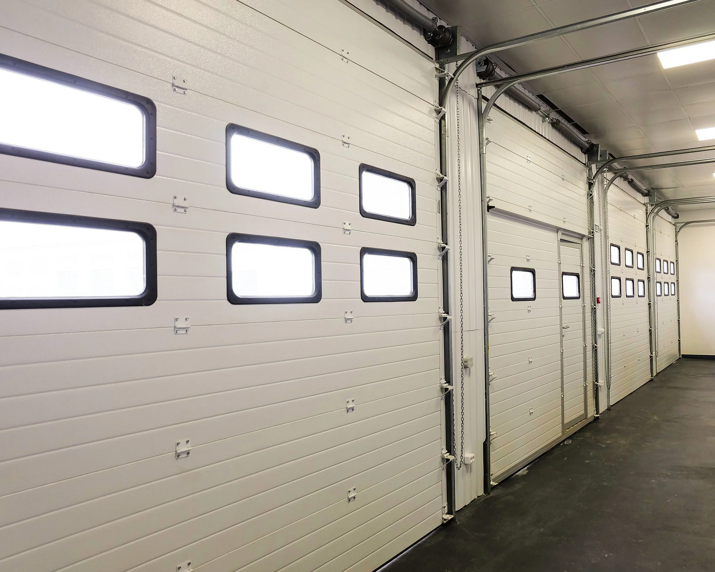 The interior of a large industrial garage with multiple white sectional overhead doors  featuring small rectangular windows at various heights, metal tracks, and automated garage door.